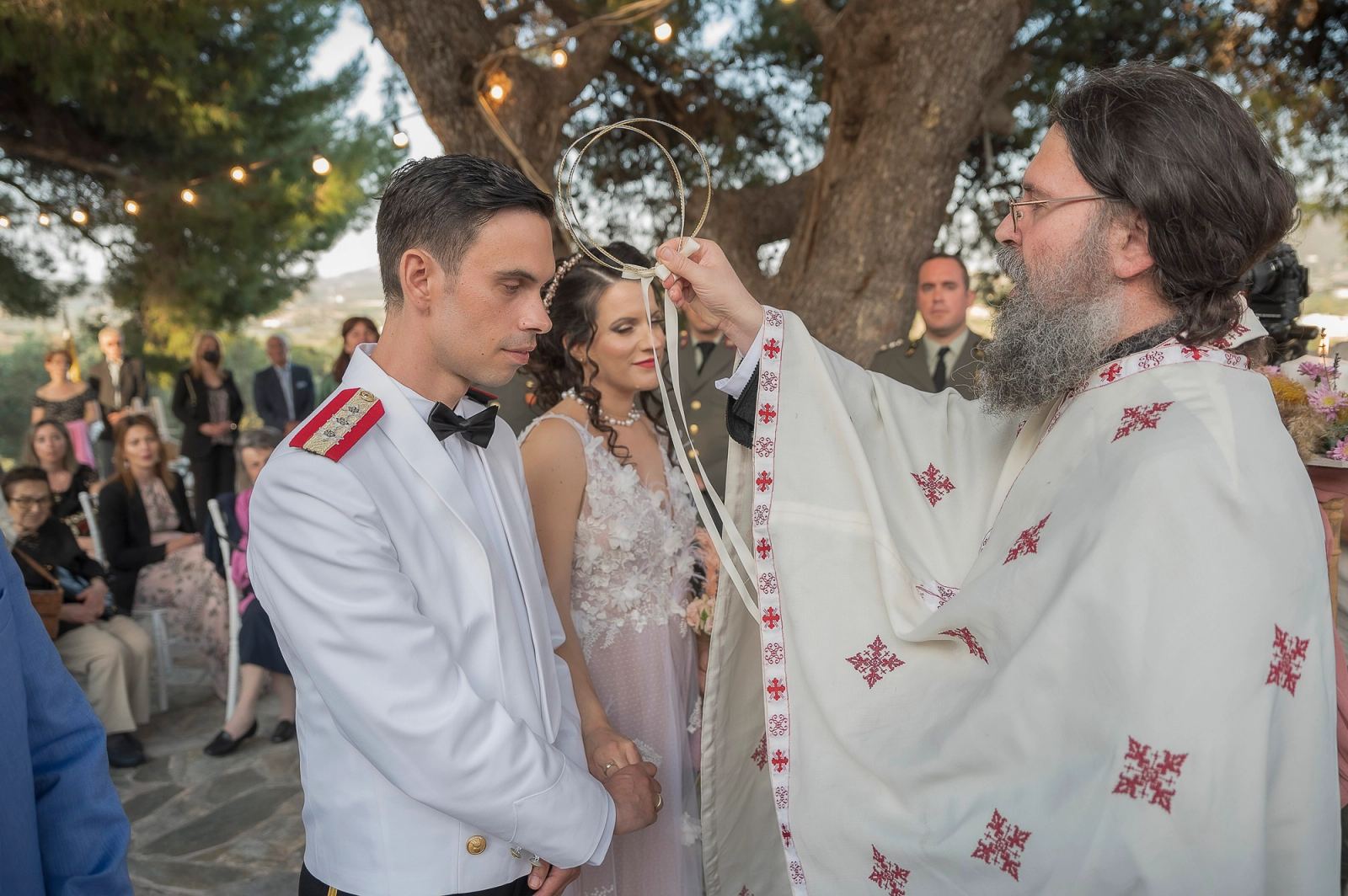 Ο ιερέας κρατά τα στέφανα πάνω από τον γαμπρό και τη νύφη σε υπαίθρια τελετή κάτω από δέντρο με φωτάκια.