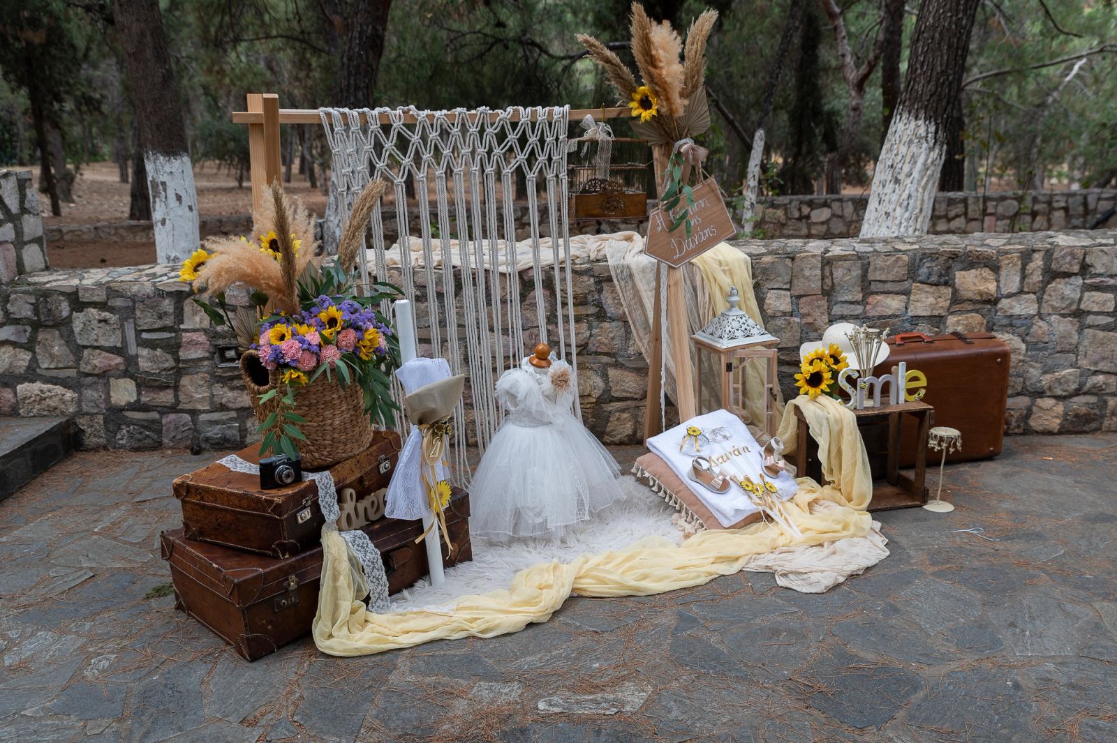 Christening setup with macramé, a dress, suitcases and sunflowers in a stone courtyard under pine trees.
