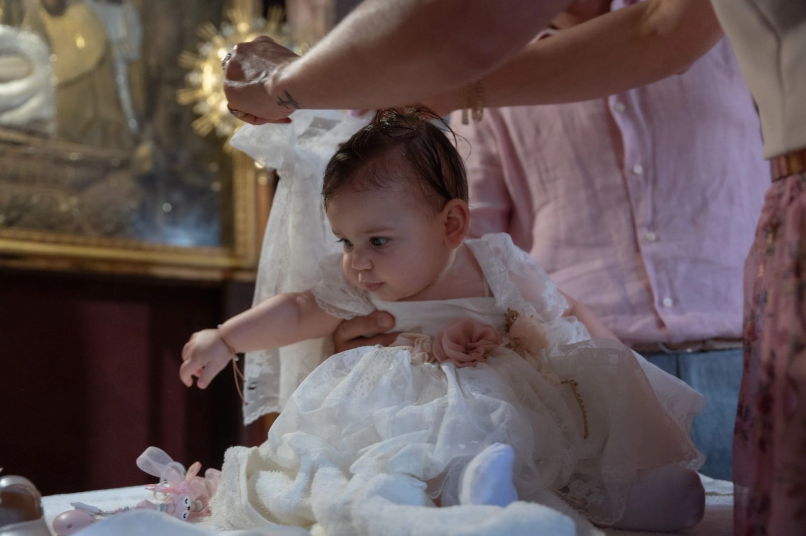 The baby with wet hair sits in a white lace dress as hands help dress her.