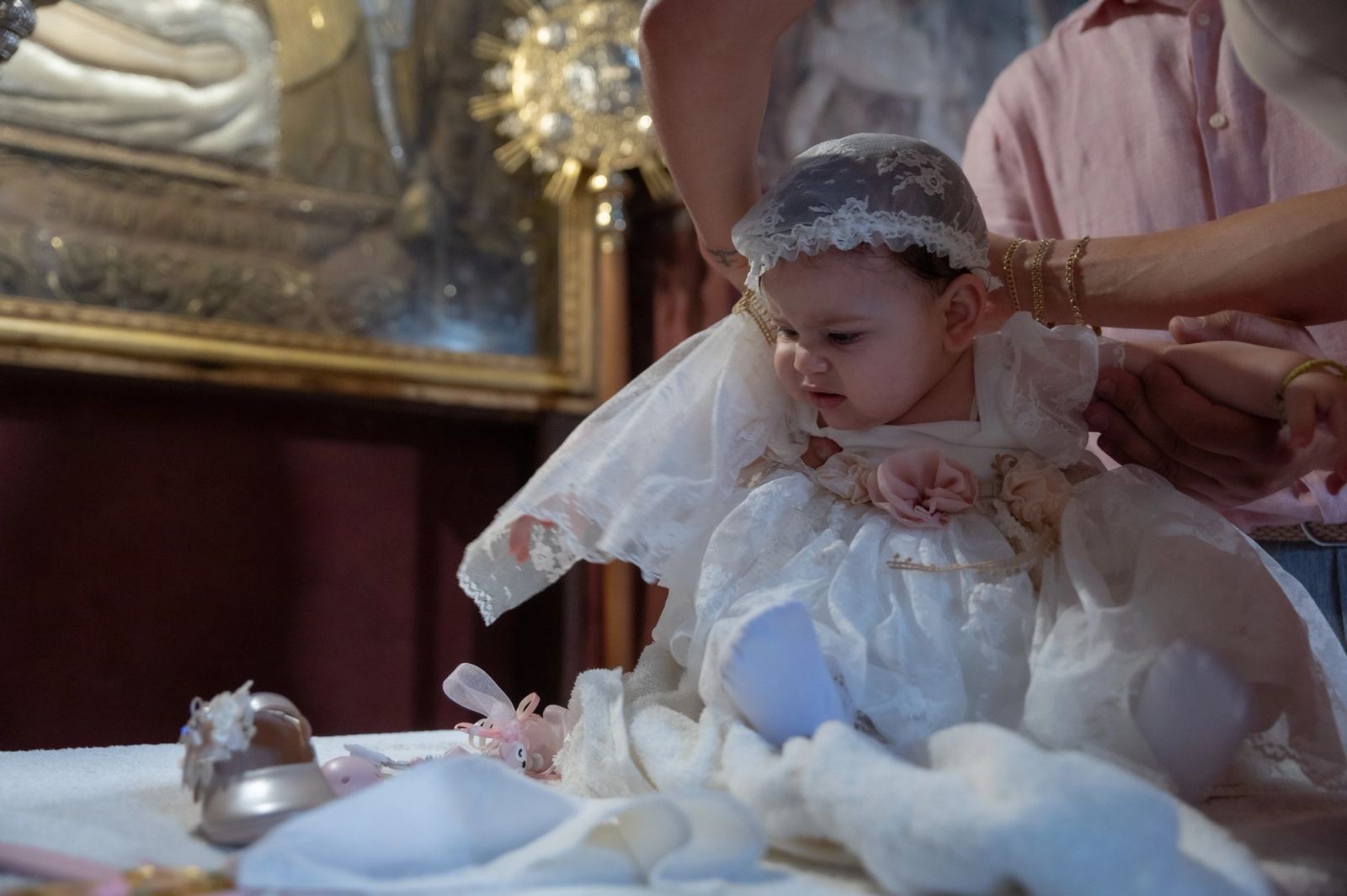 The baby in a white lace dress and bonnet sits on a table before a gold framed icon.