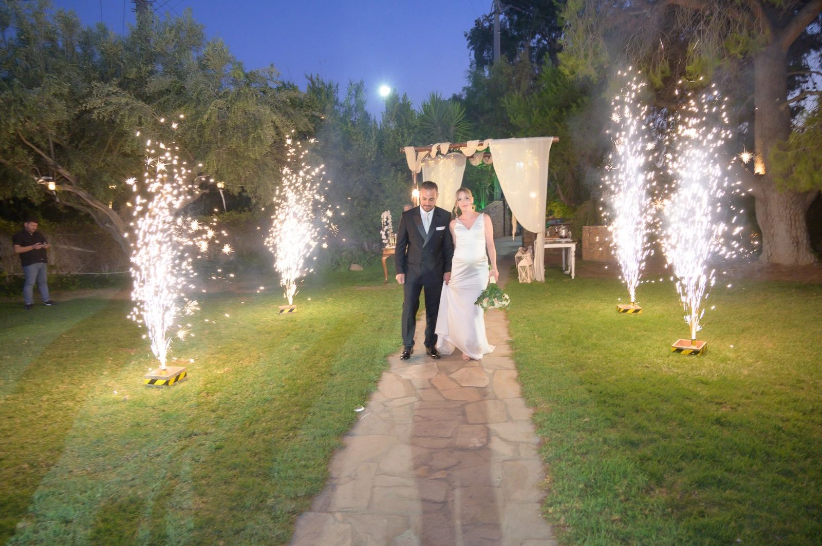 The bride and groom walk a stone path across the lawn between sparkler fountains at dusk.