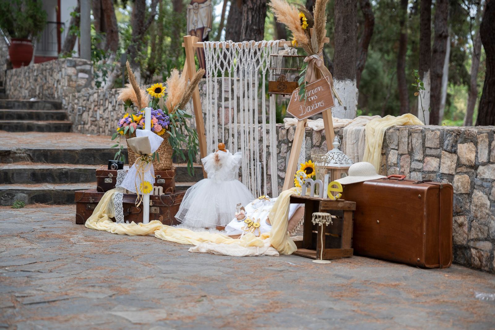 Christening decor with macramé, a white dress, a wooden sign and leather suitcases beside a stone wall.