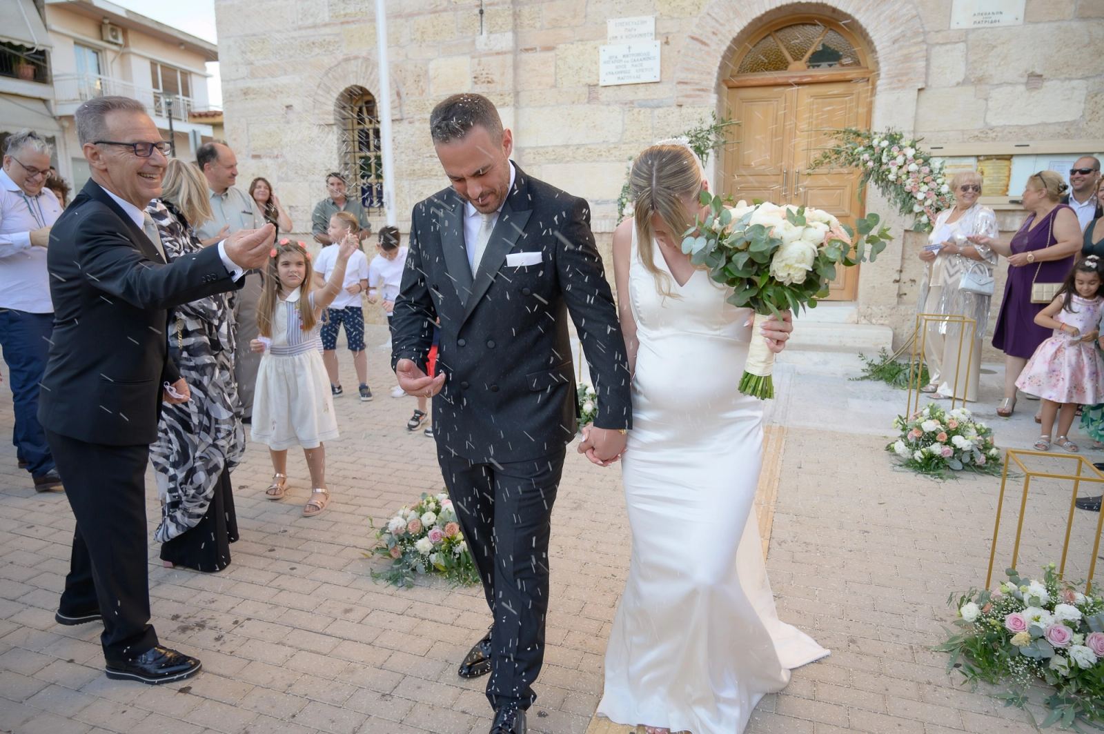 The bride and groom walk hand in hand on paving stones as guests and children shower them with rice.