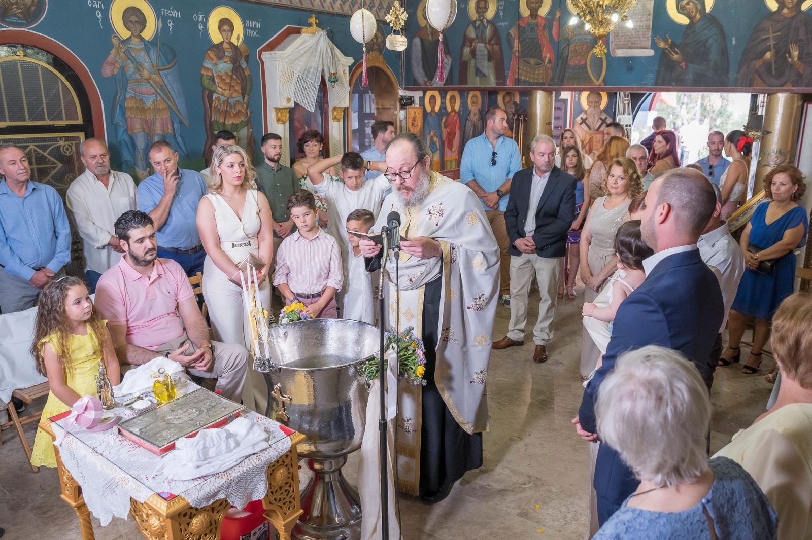 The priest reads beside the font as guests stand in a church with painted icons.
