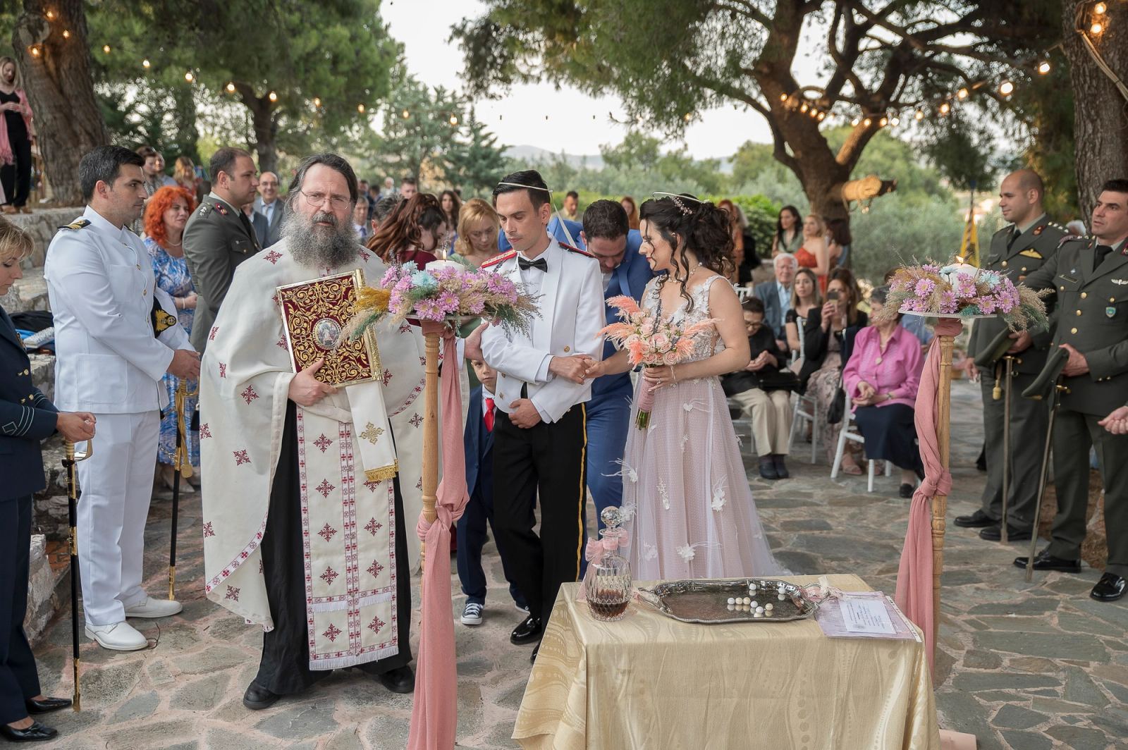Ο ιερέας κρατά Ευαγγέλιο δίπλα στη νύφη και τον γαμπρό σε πέτρινη αυλή με πεύκα και φωτάκια.