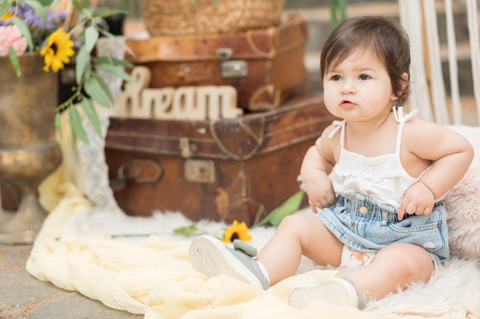 The baby sits on a fluffy rug beside vintage suitcases, sunflowers and a wooden word dream.