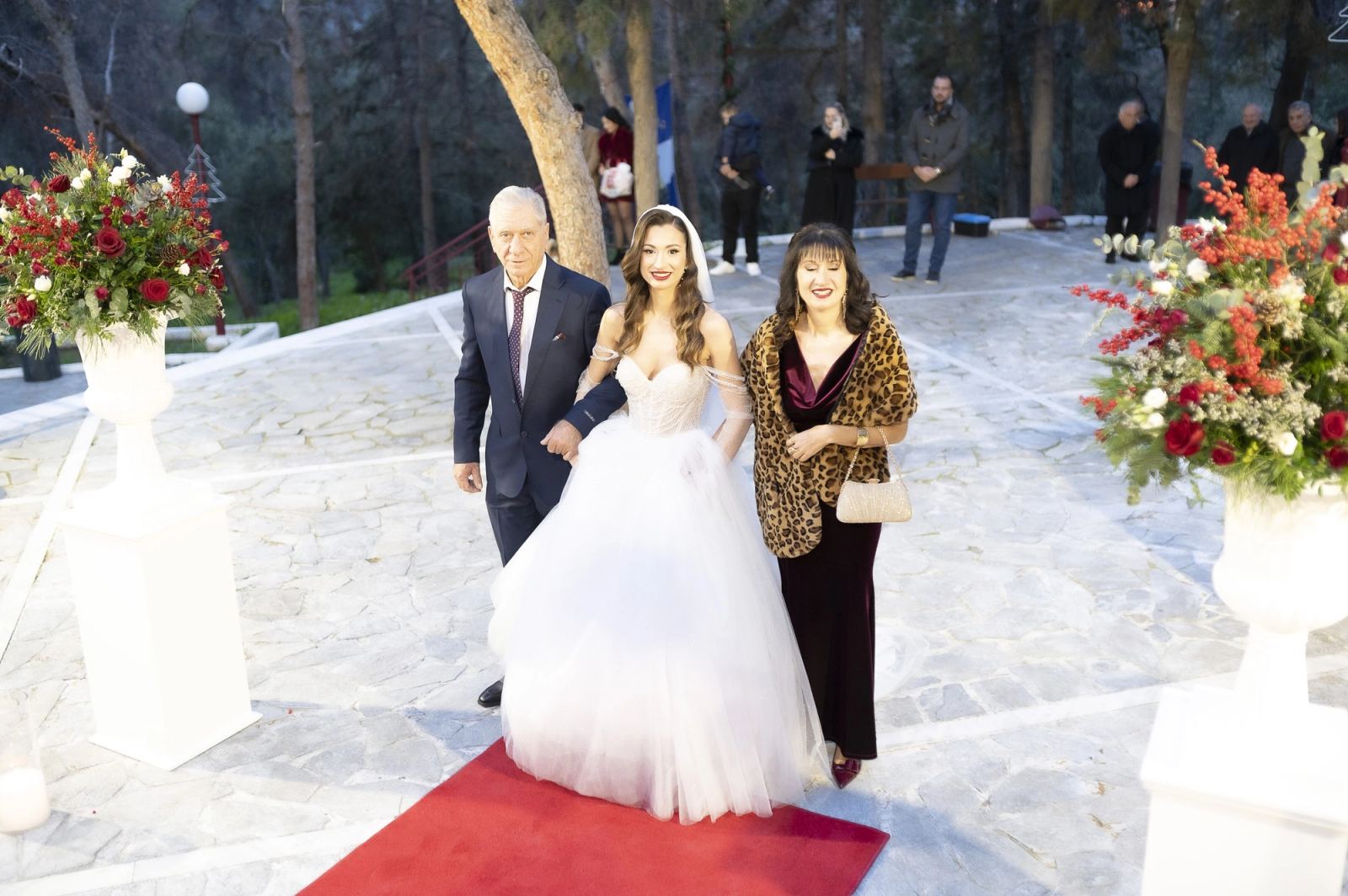 The bride walks on a red carpet with an older man and a woman in a paved courtyard