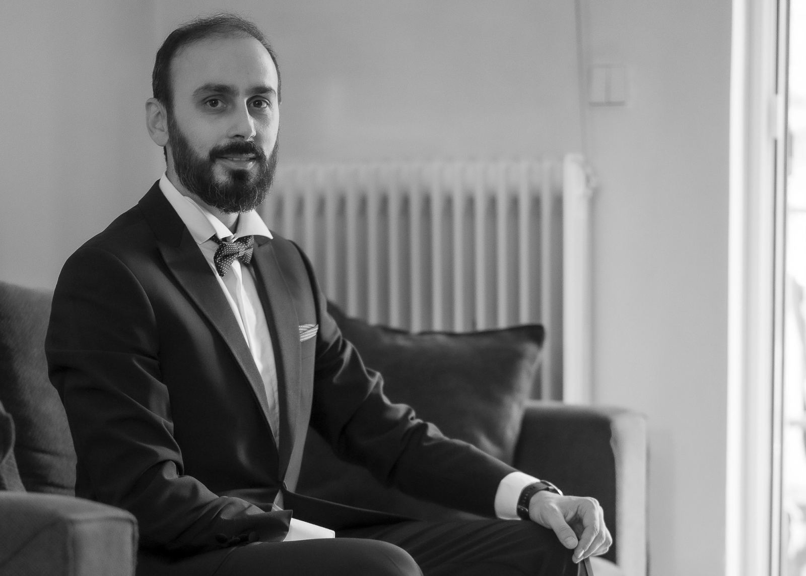 Ασπρόμαυρο πορτρέτο του γαμπρού με κοστούμι και παπιγιόν, καθισμένου σε καναπέ δίπλα σε παράθυρο.