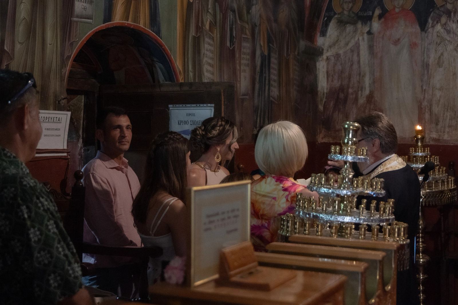 Guests sit in a church with frescoes beside candle stands as the priest stands before them.