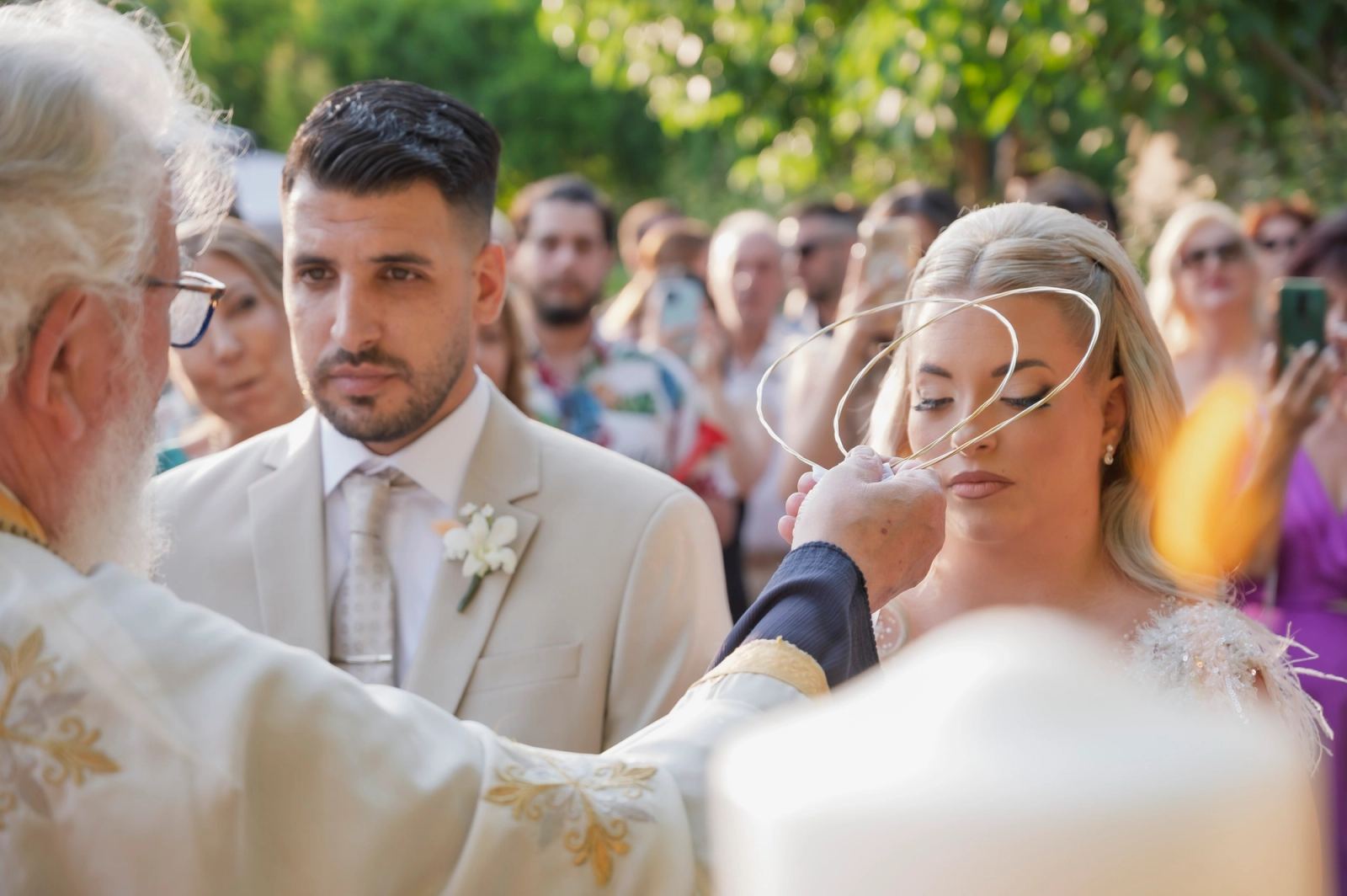 Ο ιερέας κρατά τα στέφανα πάνω από τη νύφη δίπλα στον γαμπρό, σε υπαίθριο μυστήριο με καλεσμένους.