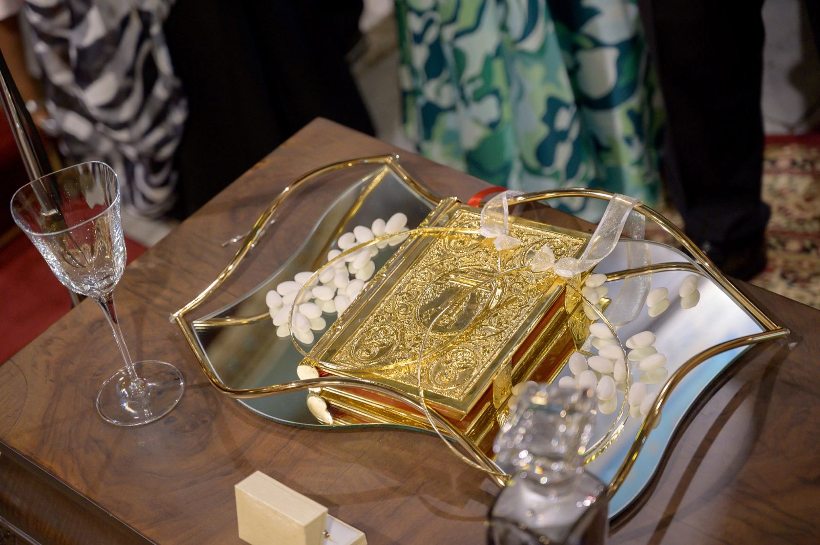 A gilded Gospel book and wedding crowns on a mirrored tray with sugared almonds, beside a crystal glass.