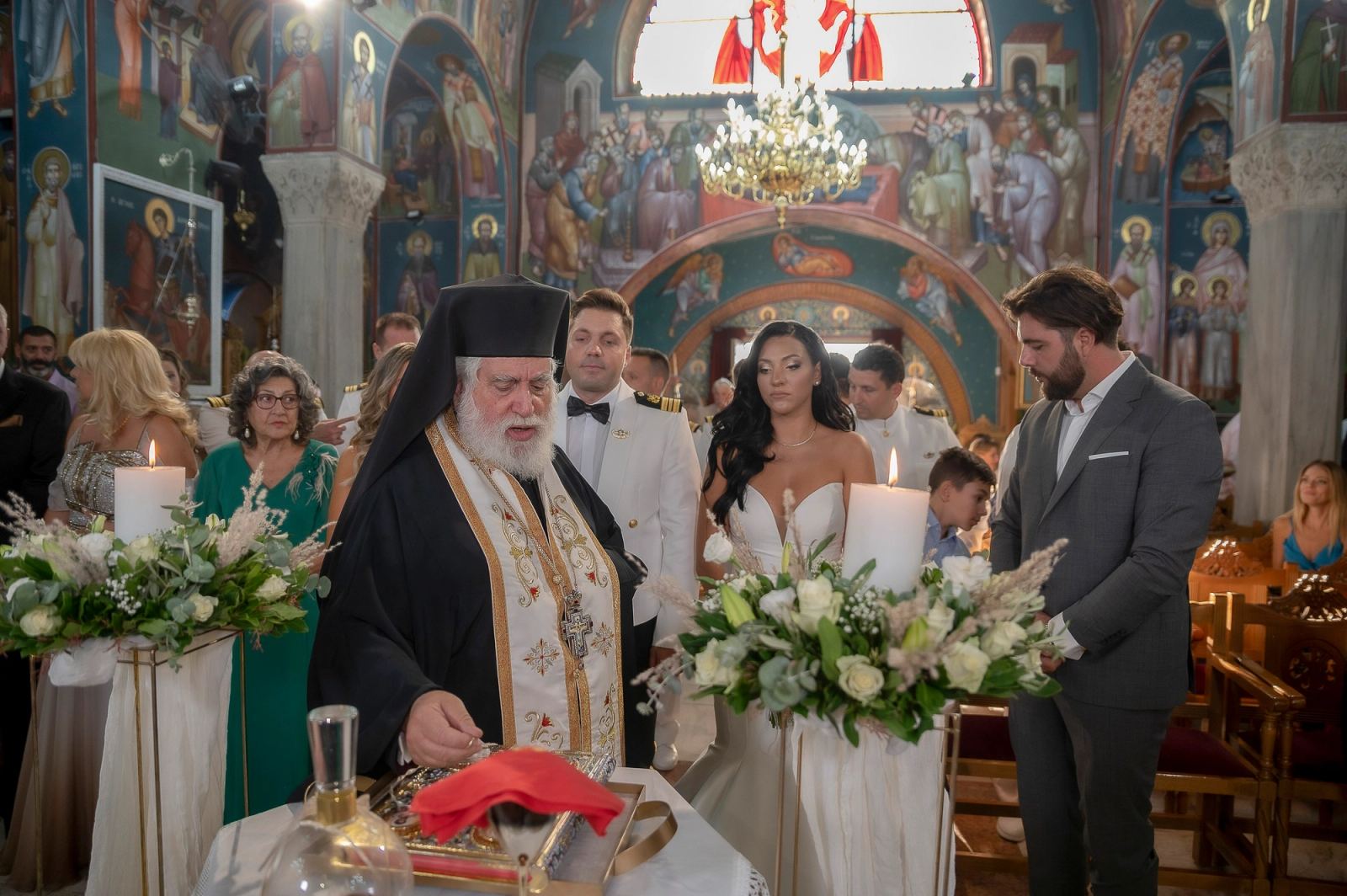 Ο ιερέας στέκεται στο τραπέζι με κρασί, ενώ το ζευγάρι και ο κουμπάρος περιμένουν δίπλα σε λαμπάδες.