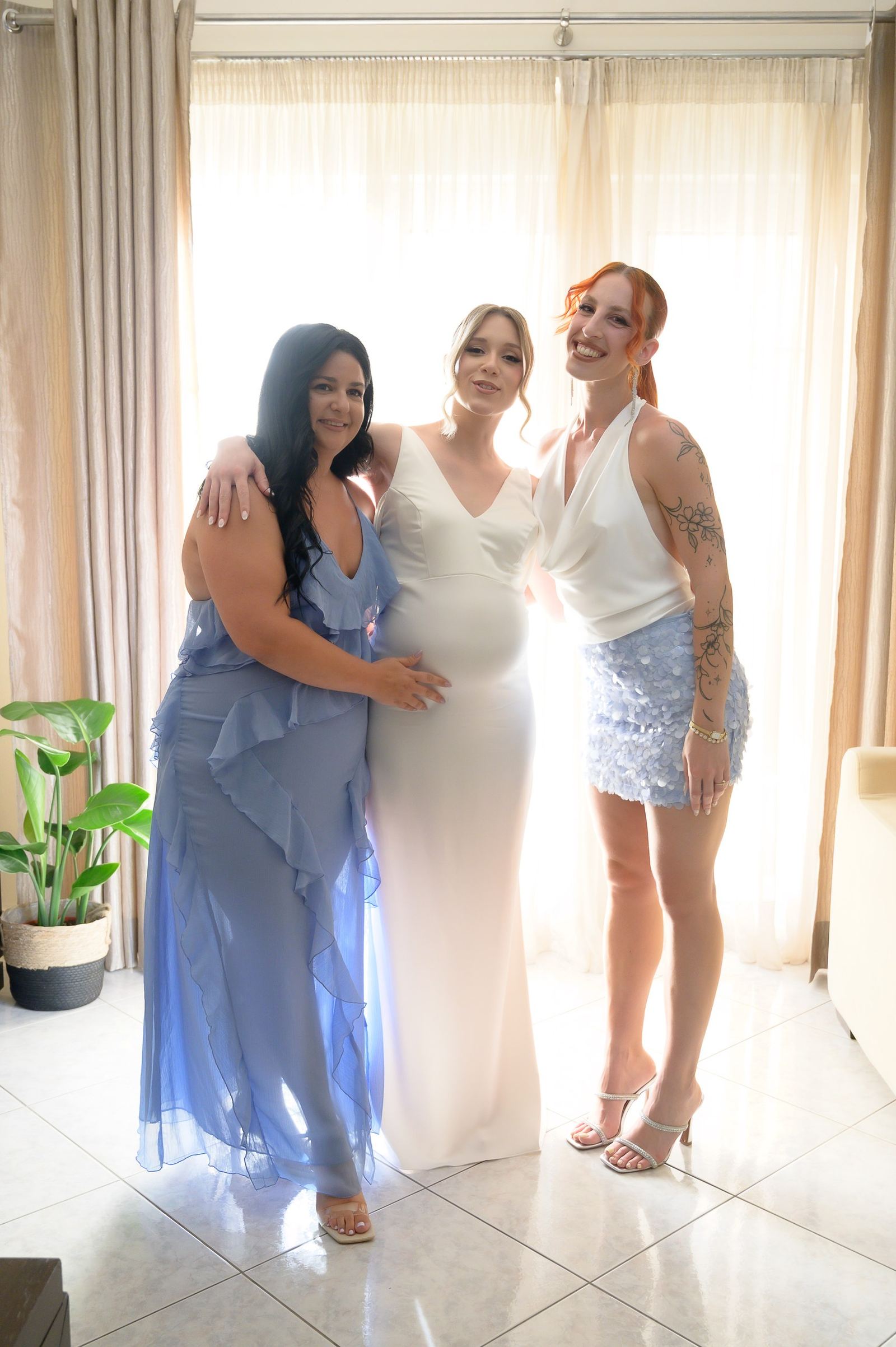 The bride in a white gown poses between two friends in front of a bright curtained window.