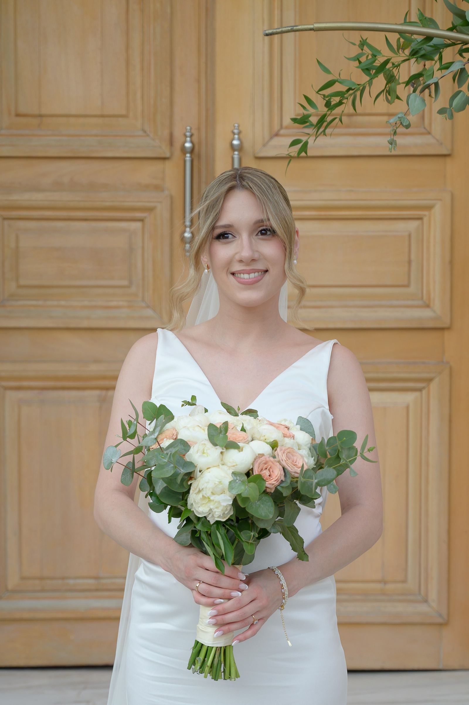 The bride smiles in front of a wooden door, holding a bouquet of white and peach flowers.