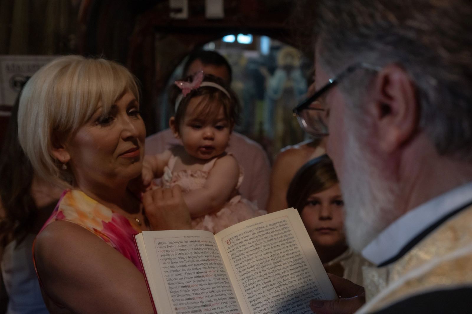 The priest in glasses reads from a book before a woman holding the baby, with a boy behind.