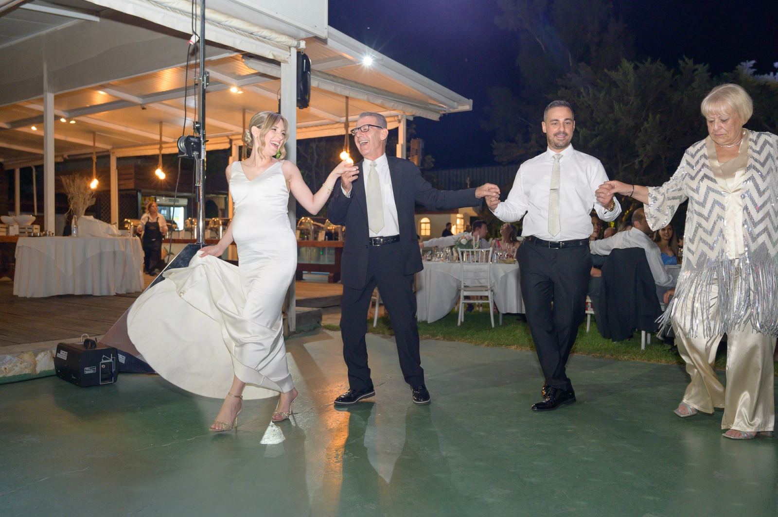 The bride laughs as she dances hand in hand with the groom and two others on an outdoor floor at night.