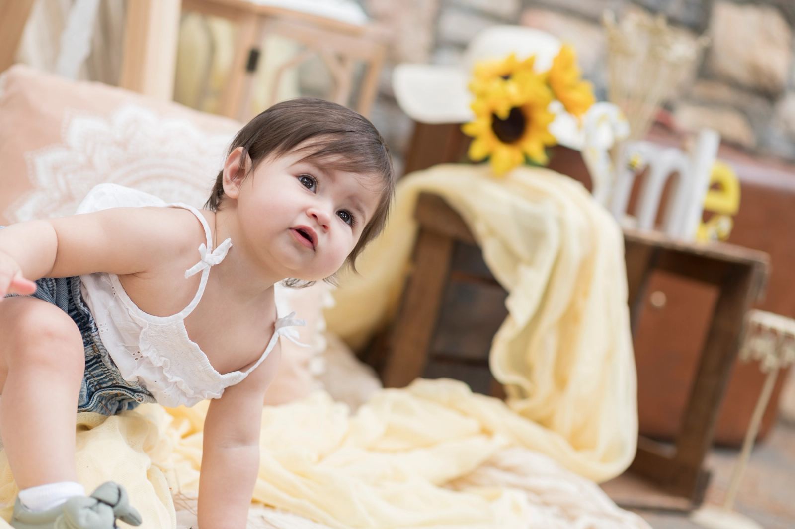 The baby crawls on a yellow cloth, with sunflowers and wooden furniture behind.