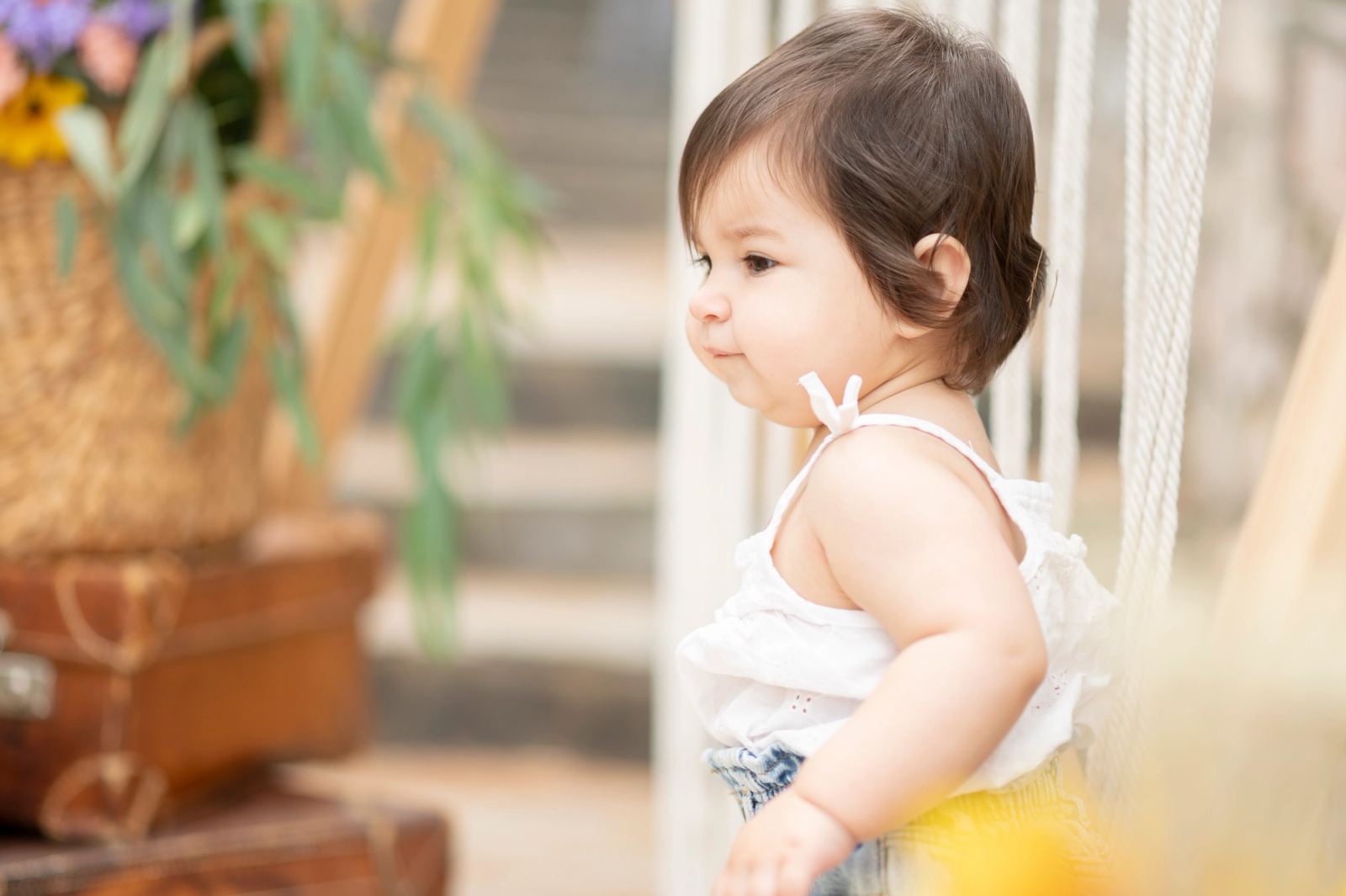 The baby in a white top in profile, before vintage suitcases and a wicker basket of flowers.