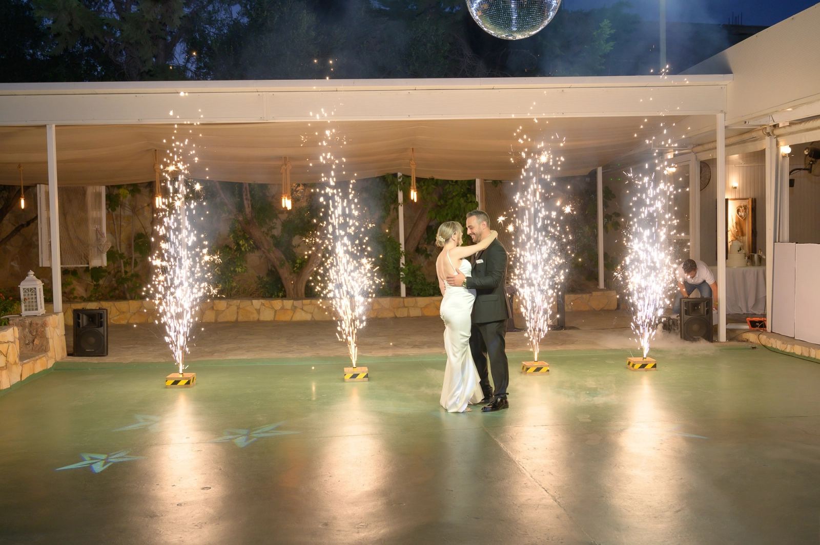 The bride and groom dance in an embrace on a dance floor with sparkler fountains under a disco ball.