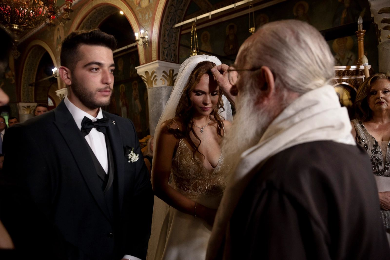 Ο ιερέας αγγίζει το μέτωπο της νύφης με πέπλο, ενώ ο γαμπρός με παπιγιόν στέκεται δίπλα της στην εκκλησία.