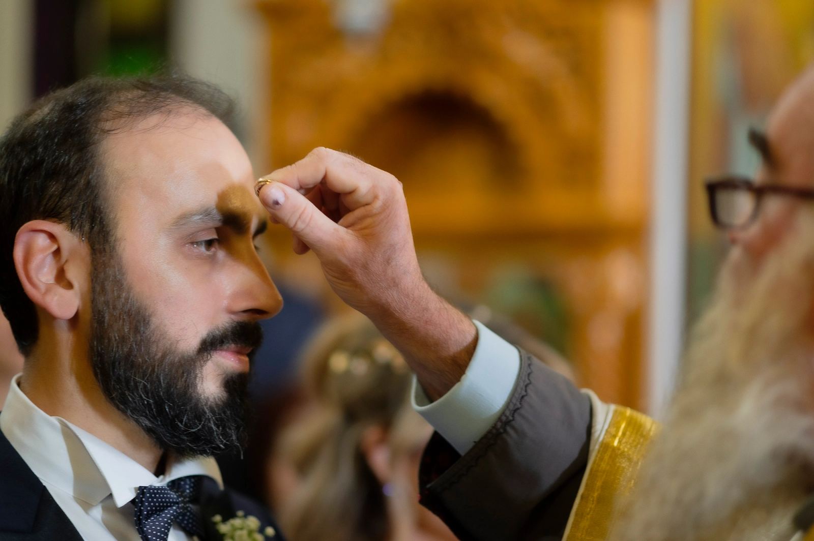 Ο ιερέας σταυρώνει με τη βέρα το μέτωπο του γαμπρού μπροστά σε χρυσό ξυλόγλυπτο τέμπλο.