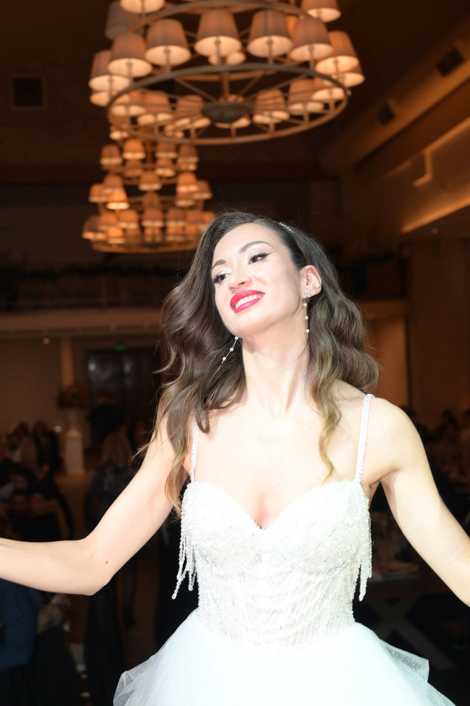 The bride with red lipstick dances with open arms under chandeliers with lampshades.