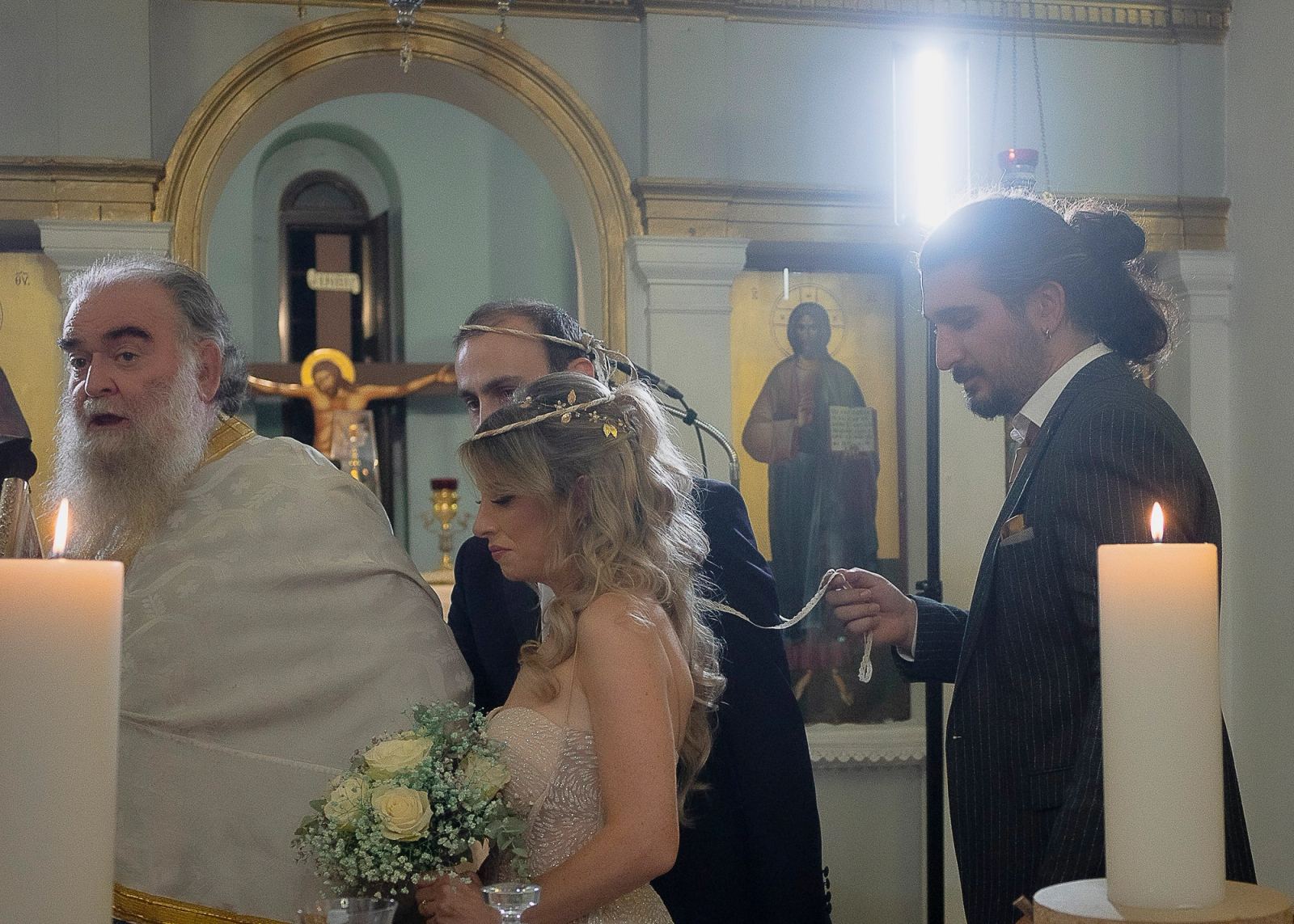Ο κουμπάρος αλλάζει τα στέφανα του ζευγαριού δίπλα στον ιερέα, ανάμεσα σε ψηλές λευκές λαμπάδες.