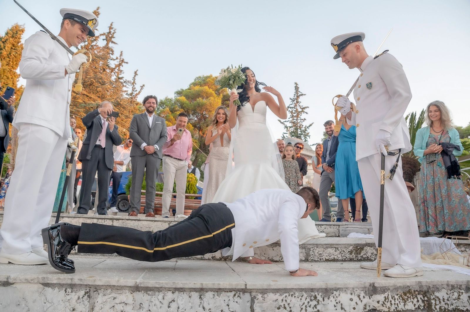 Ο γαμπρός κάνει κάμψεις στα πέτρινα σκαλιά μπροστά στη νύφη, ενώ ναυτικοί με σπαθιά και καλεσμένοι κοιτούν.
