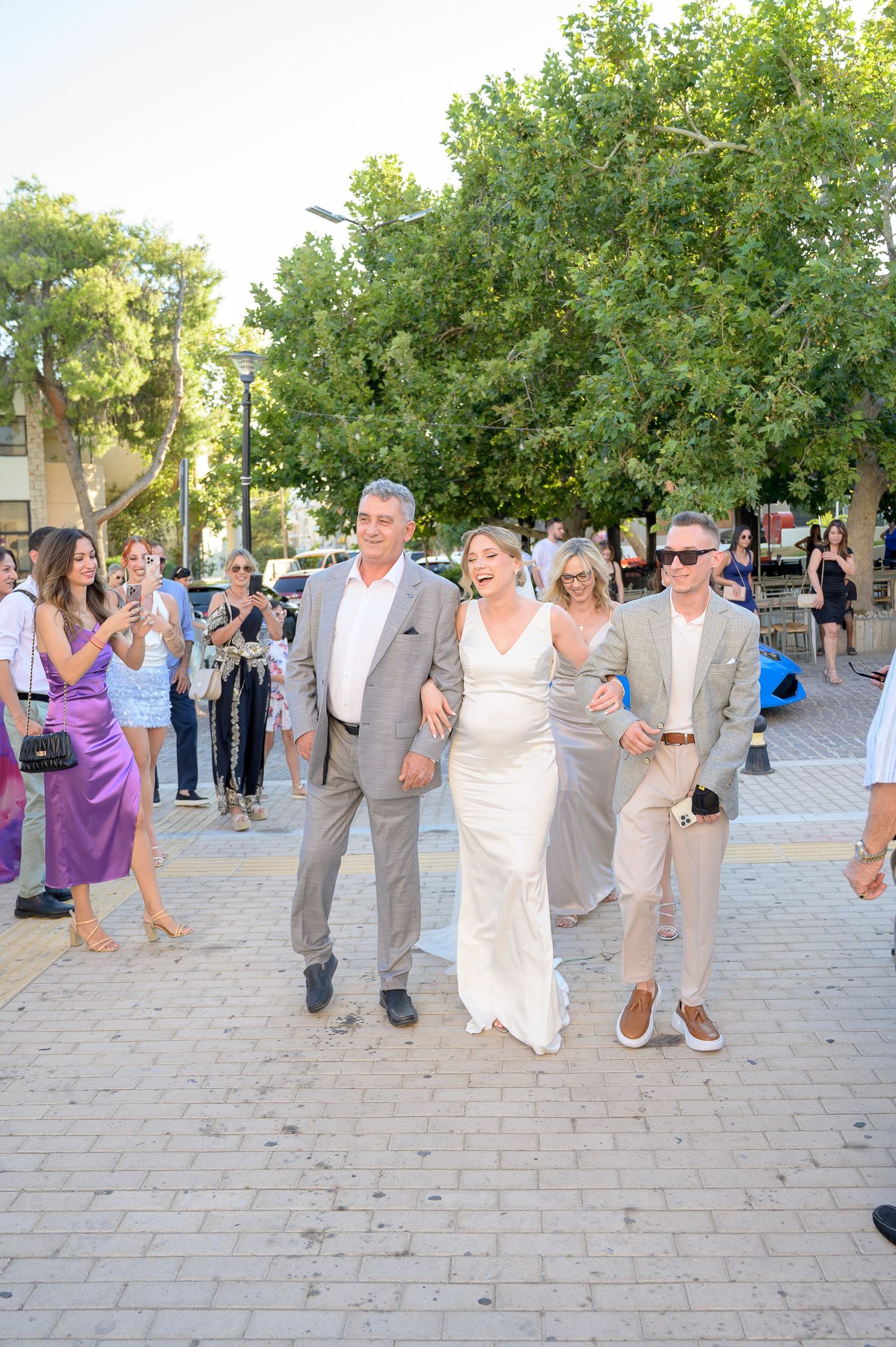 The bride walks across a square arm in arm with two men in grey jackets as guests take photos.