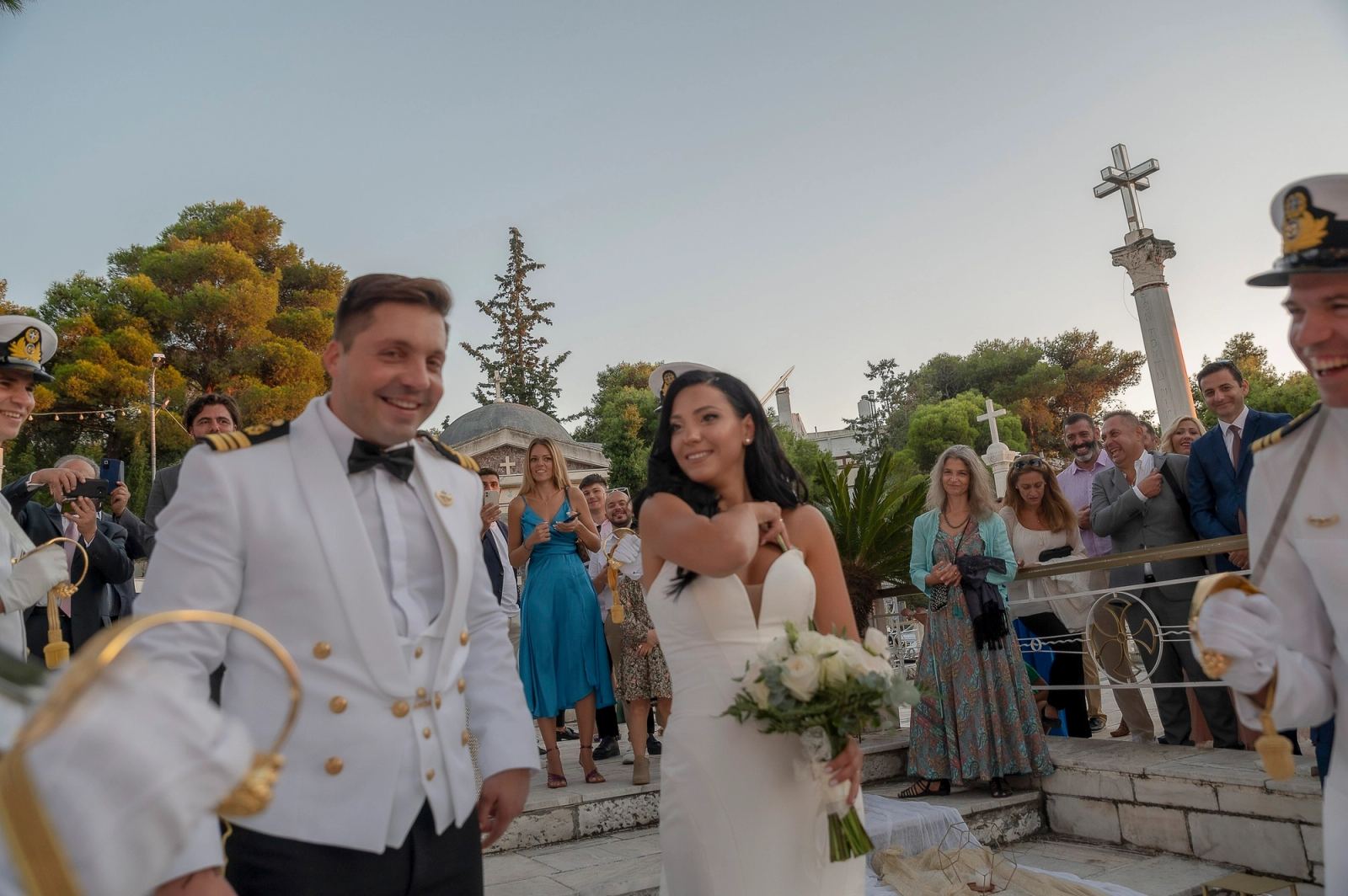 Η νύφη και ο γαμπρός χαμογελούν σε πλατεία με καλεσμένους, δέντρα και ψηλό πέτρινο σταυρό στο βάθος.