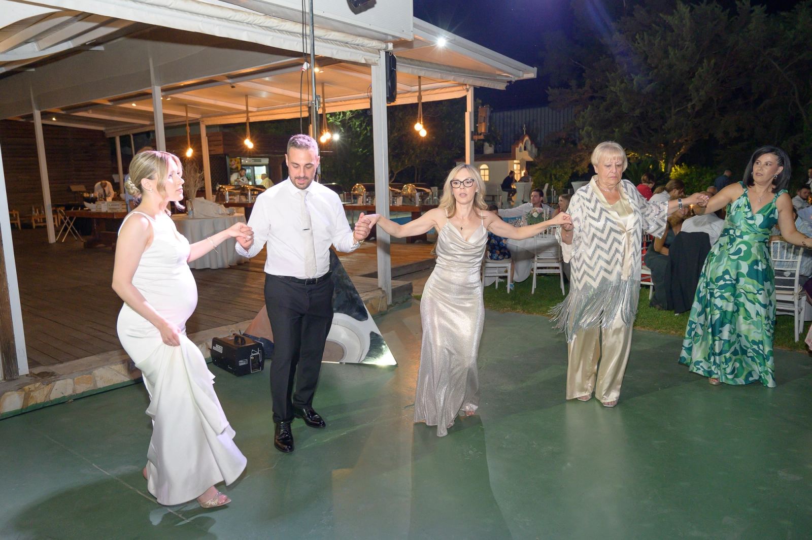 The bride, the groom and female guests dance holding hands on an outdoor dance floor at night.