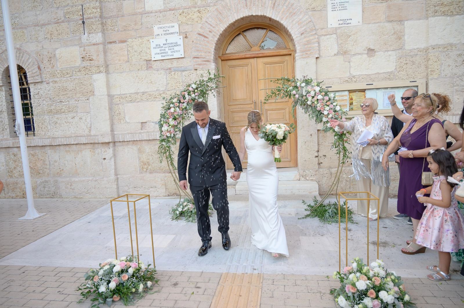 The bride and groom leave the Church of the Analipsi under a floral arch as guests throw rice.