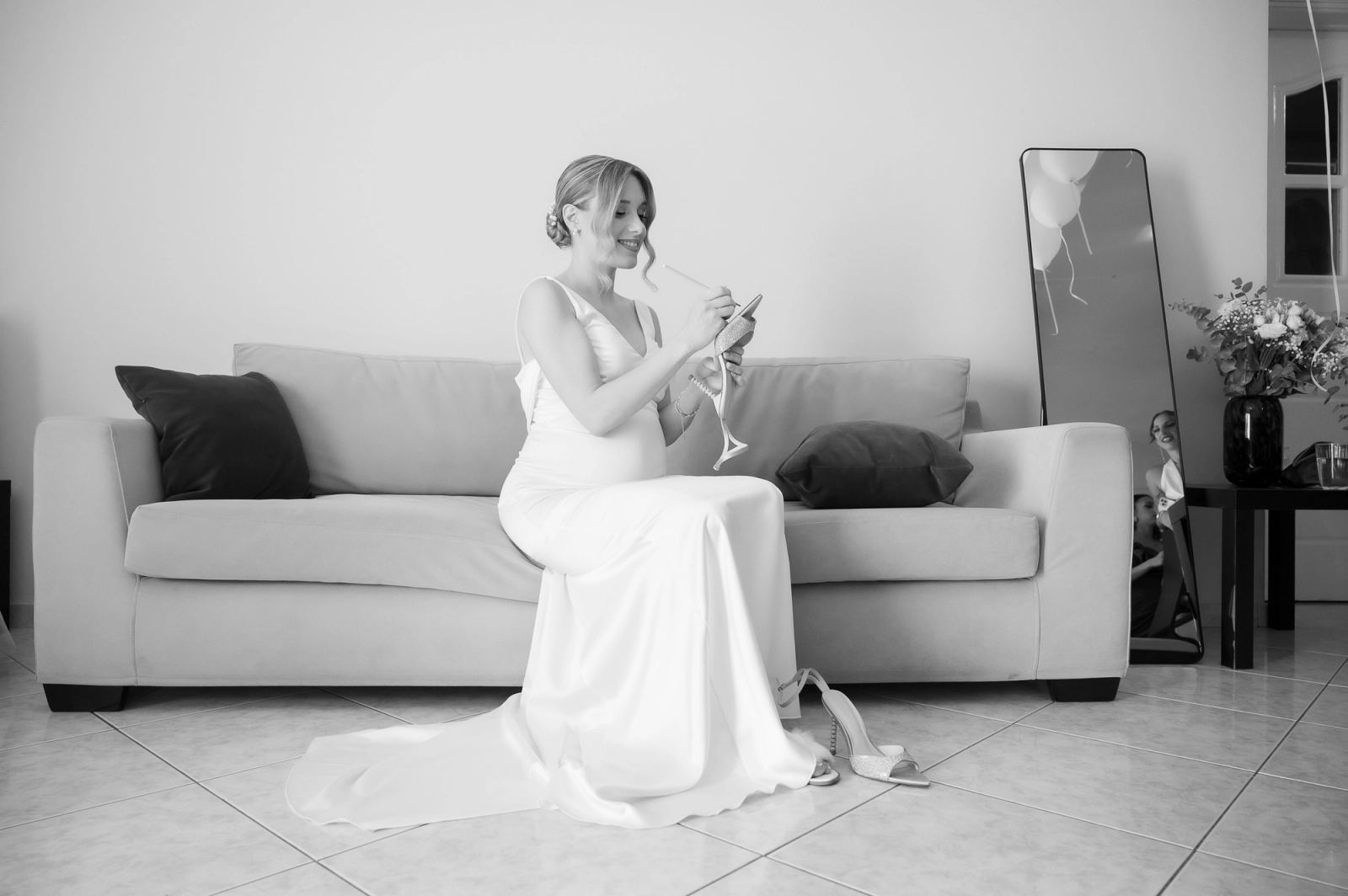 The bride in a white satin gown sits on a sofa writing in a notebook, her heels on the floor beside her.