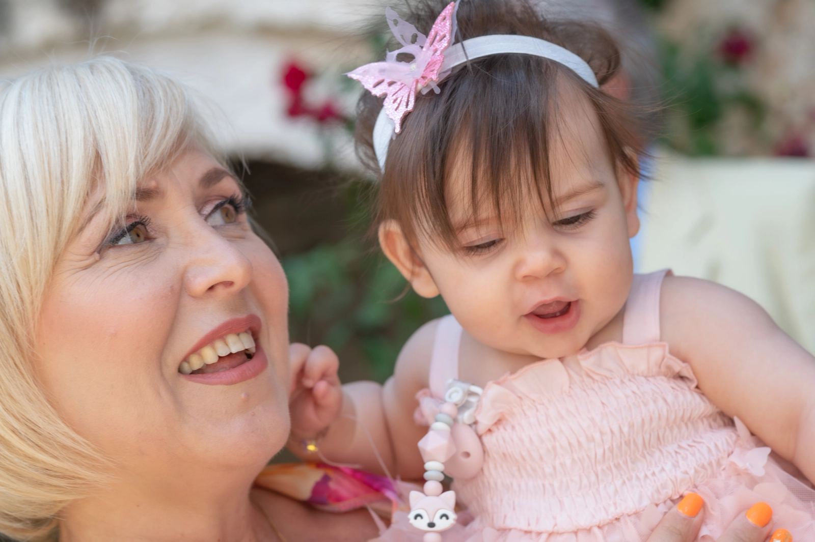 A blonde woman smiles beside the baby in a pink dress and butterfly headband.