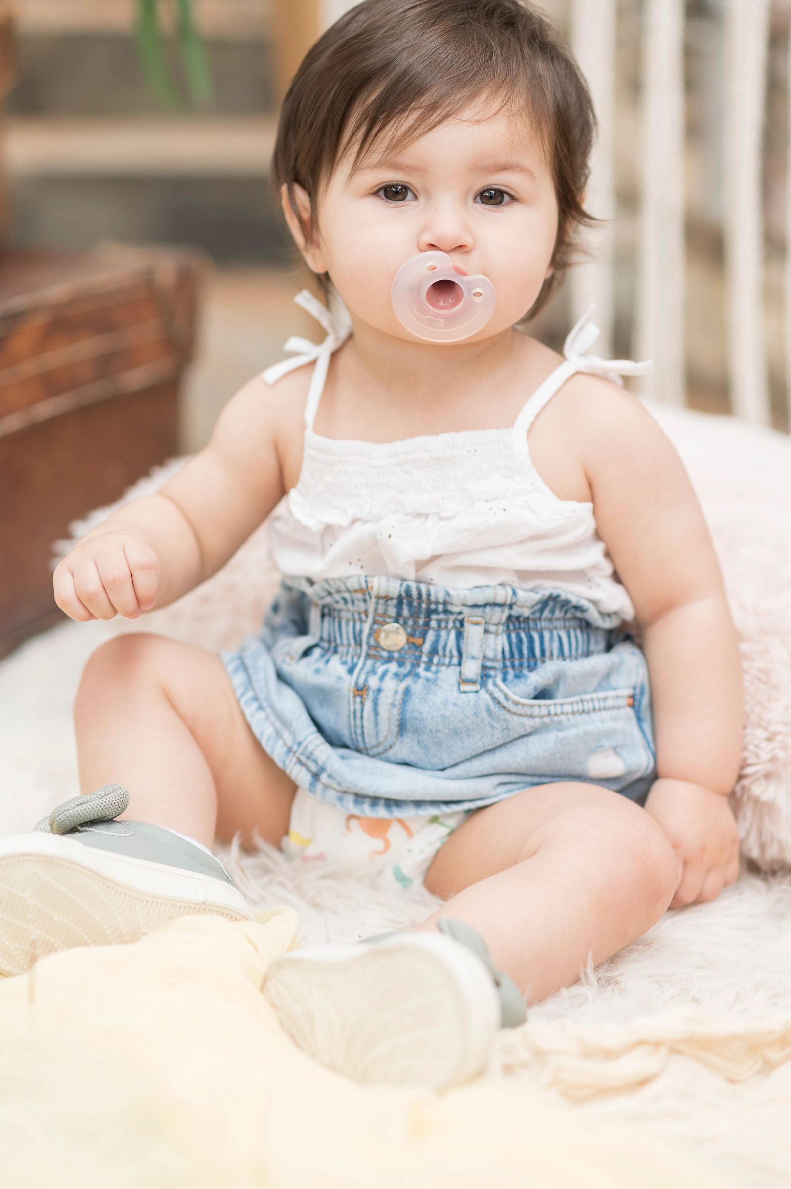 The baby in a white top, denim skirt and pacifier sits on a white fluffy rug.
