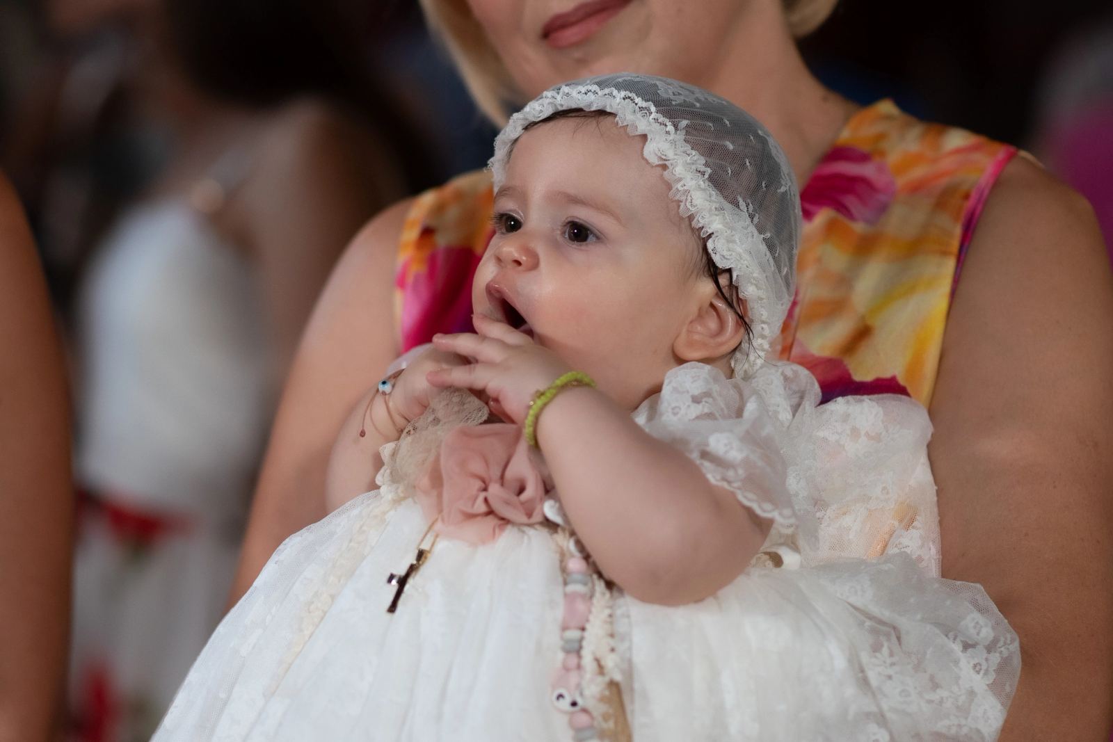 The baby in a white lace dress and bonnet puts a hand to her mouth in a woman's arms.