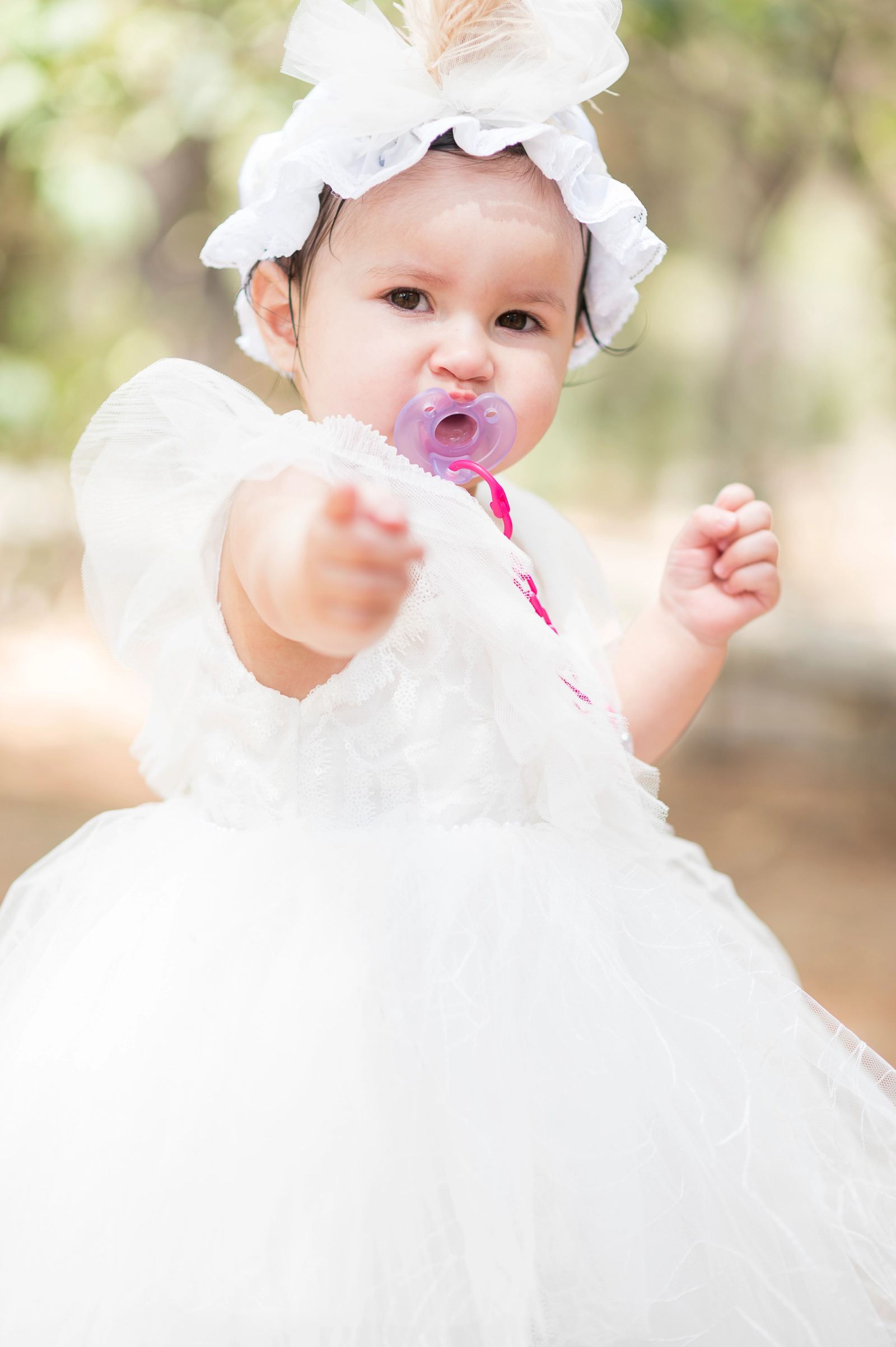 The baby in a white tulle dress, hair bow and pink pacifier reaches out a hand.