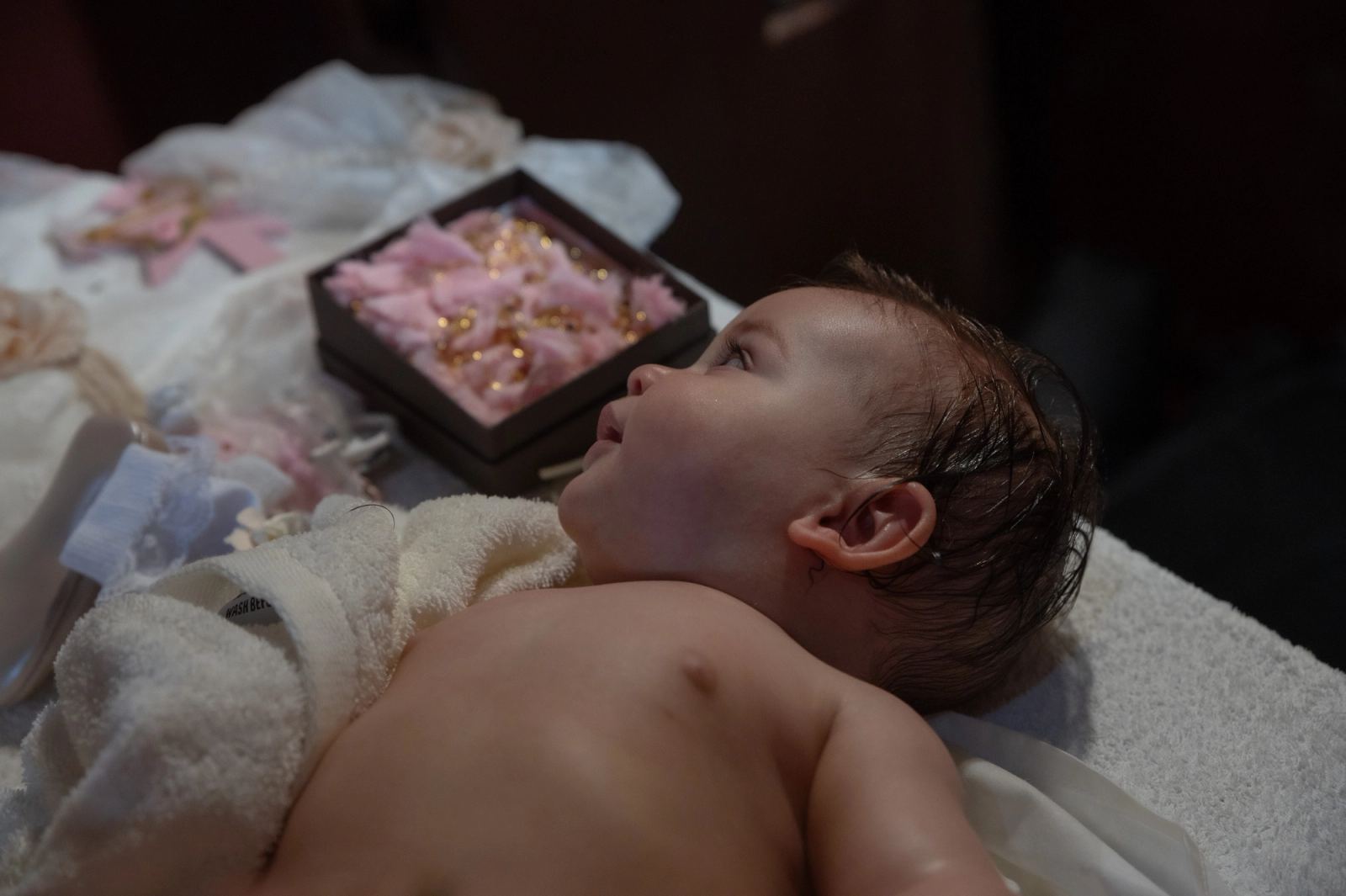 The baby lies on a white towel looking up, beside a box of pink favors.