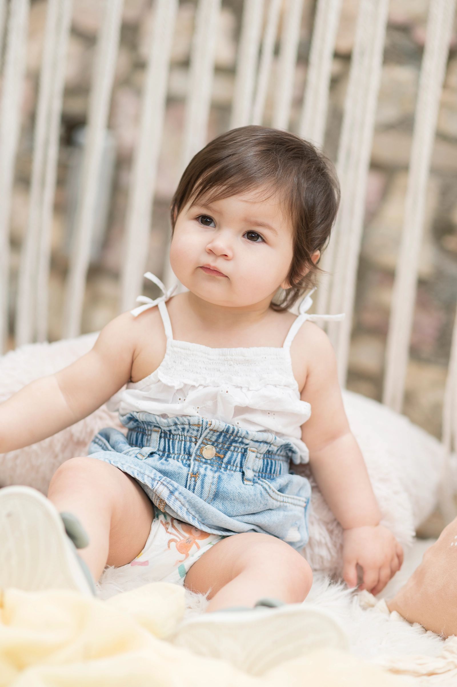The baby in a white top and denim skirt sits on a fluffy rug in front of macramé fringes.