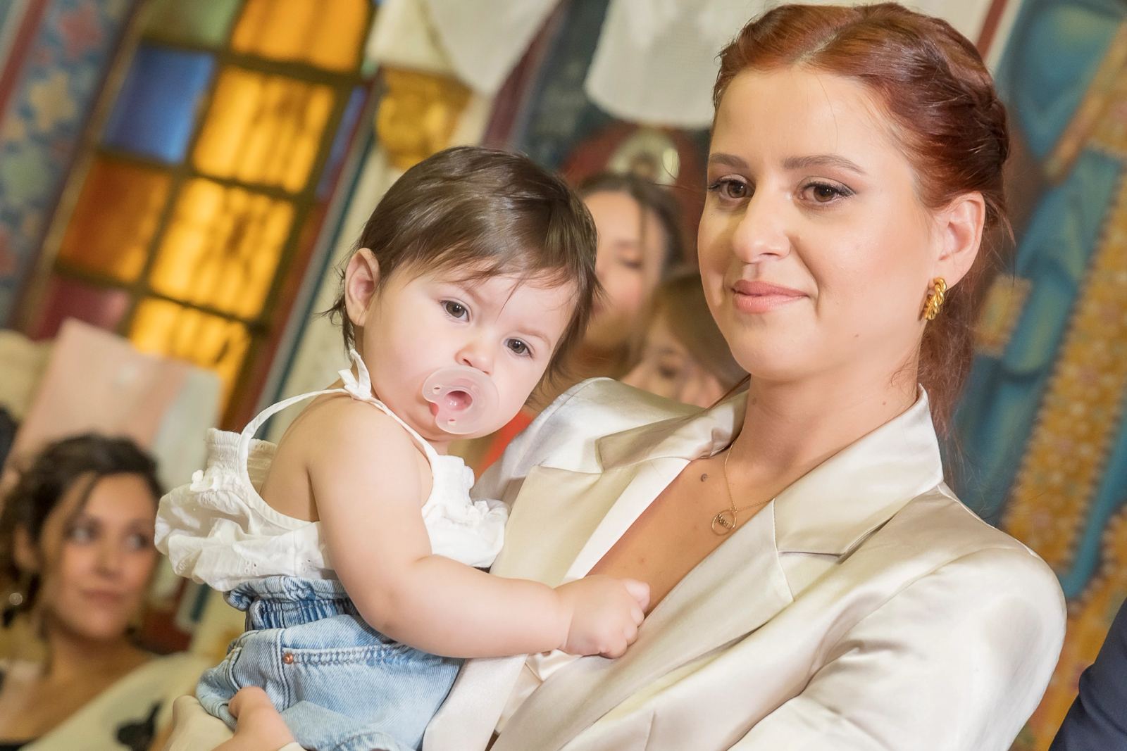 A woman in a white blazer holds the baby with a pacifier inside a church with stained glass.