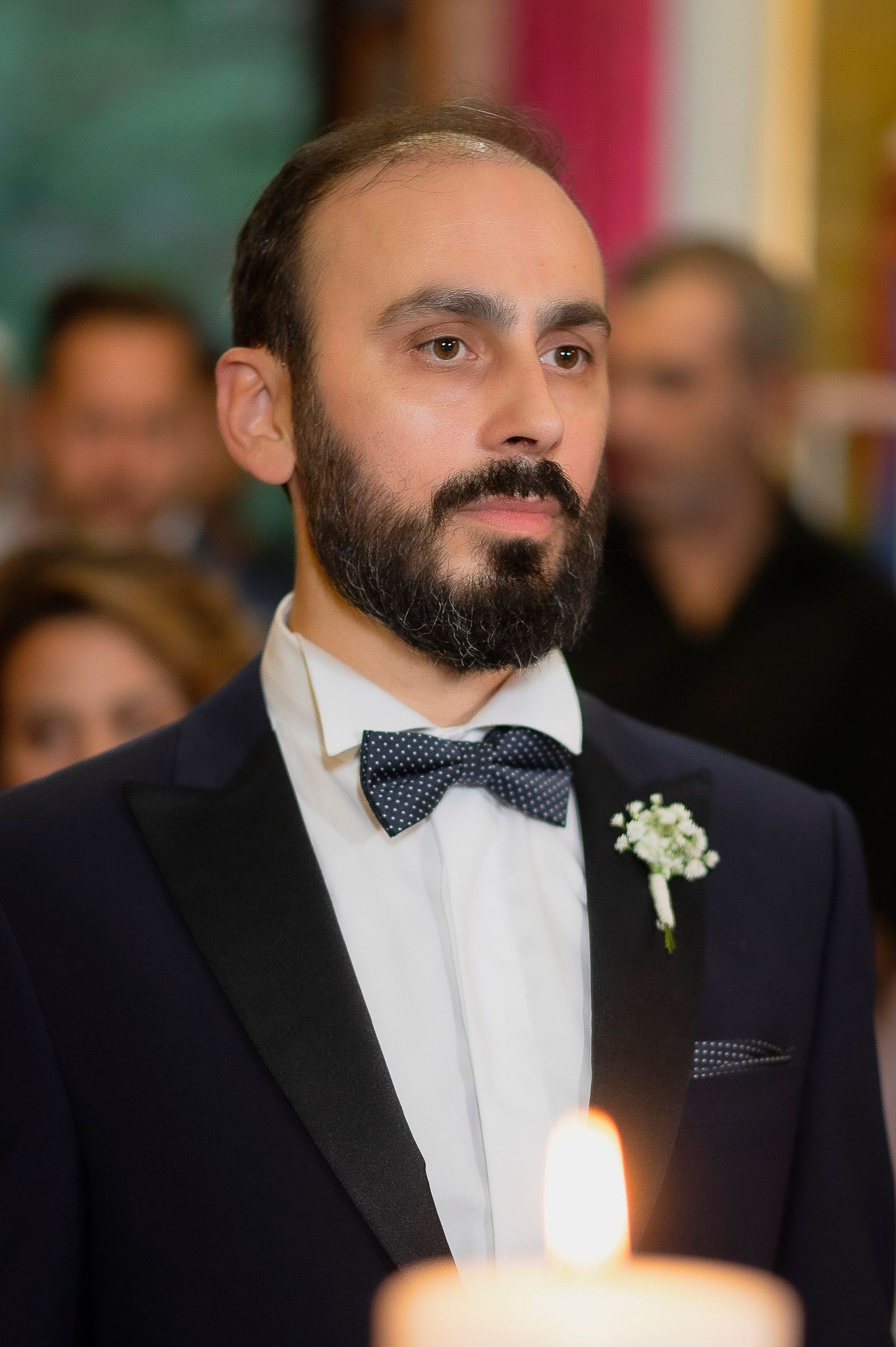 Ο γαμπρός με σμόκιν, παπιγιόν με πουά και λευκά λουλούδια στο πέτο, πίσω από αναμμένο κερί.