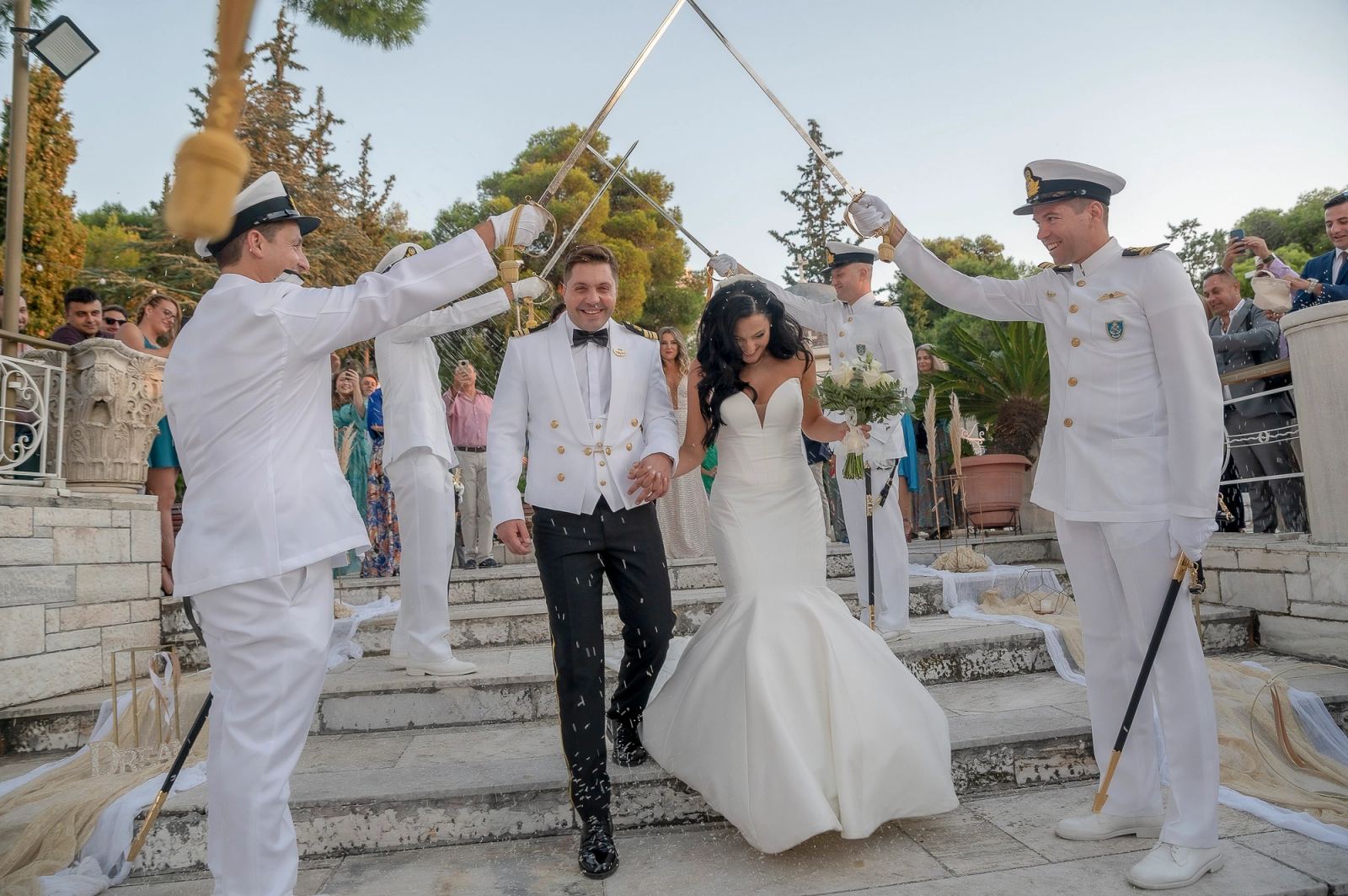 Η νύφη και ο γαμπρός κατεβαίνουν πέτρινα σκαλιά κάτω από αψίδα από σπαθιά ναυτικών με λευκές στολές.