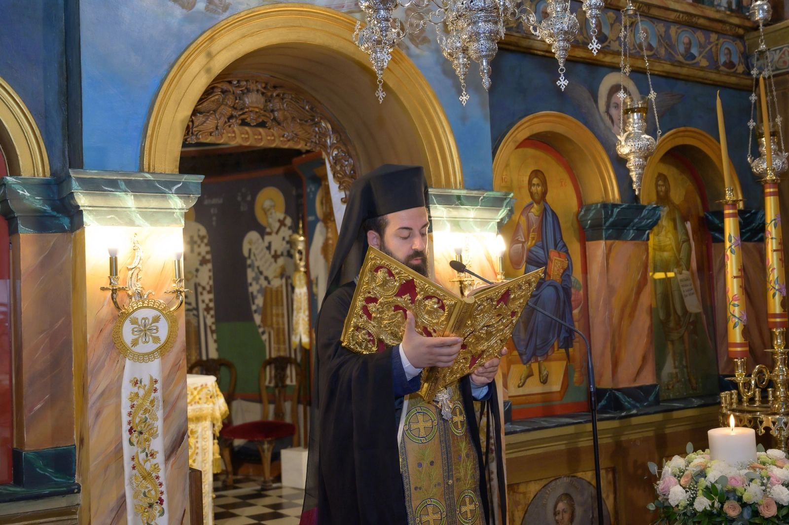 The priest reads from a gilded book before an iconostasis with painted icons and lit candles.
