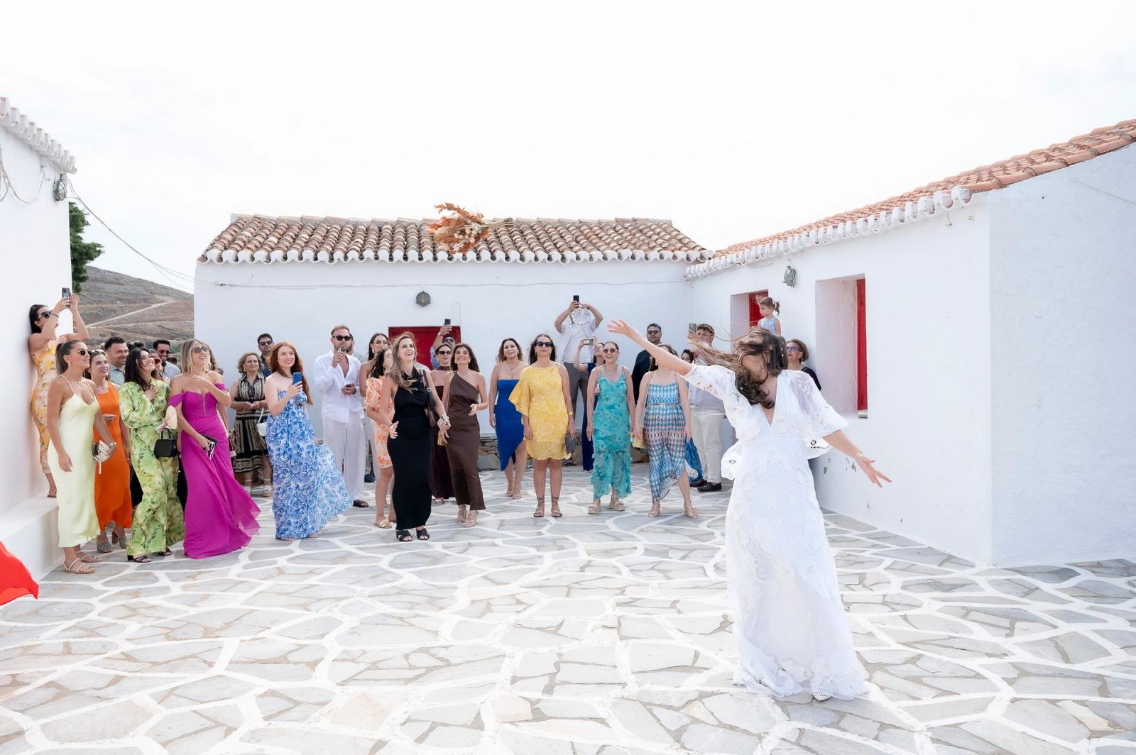 Η νύφη πετά την ανθοδέσμη πάνω από το κεφάλι της προς τις καλεσμένες σε λευκή πλακόστρωτη αυλή.