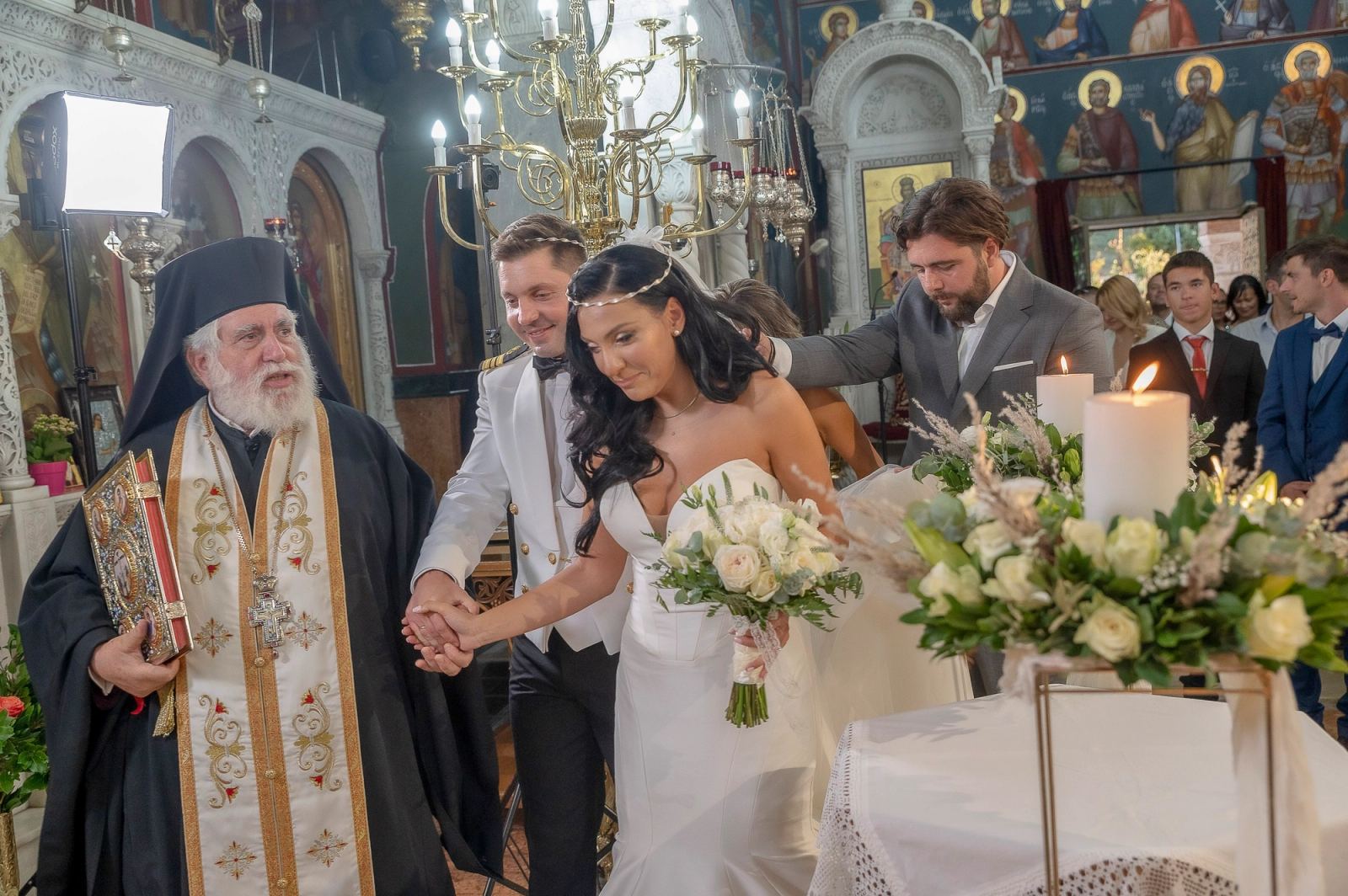 Ο ιερέας οδηγεί το ζευγάρι γύρω από το τραπέζι, με τον κουμπάρο πίσω τους και αναμμένες λαμπάδες.