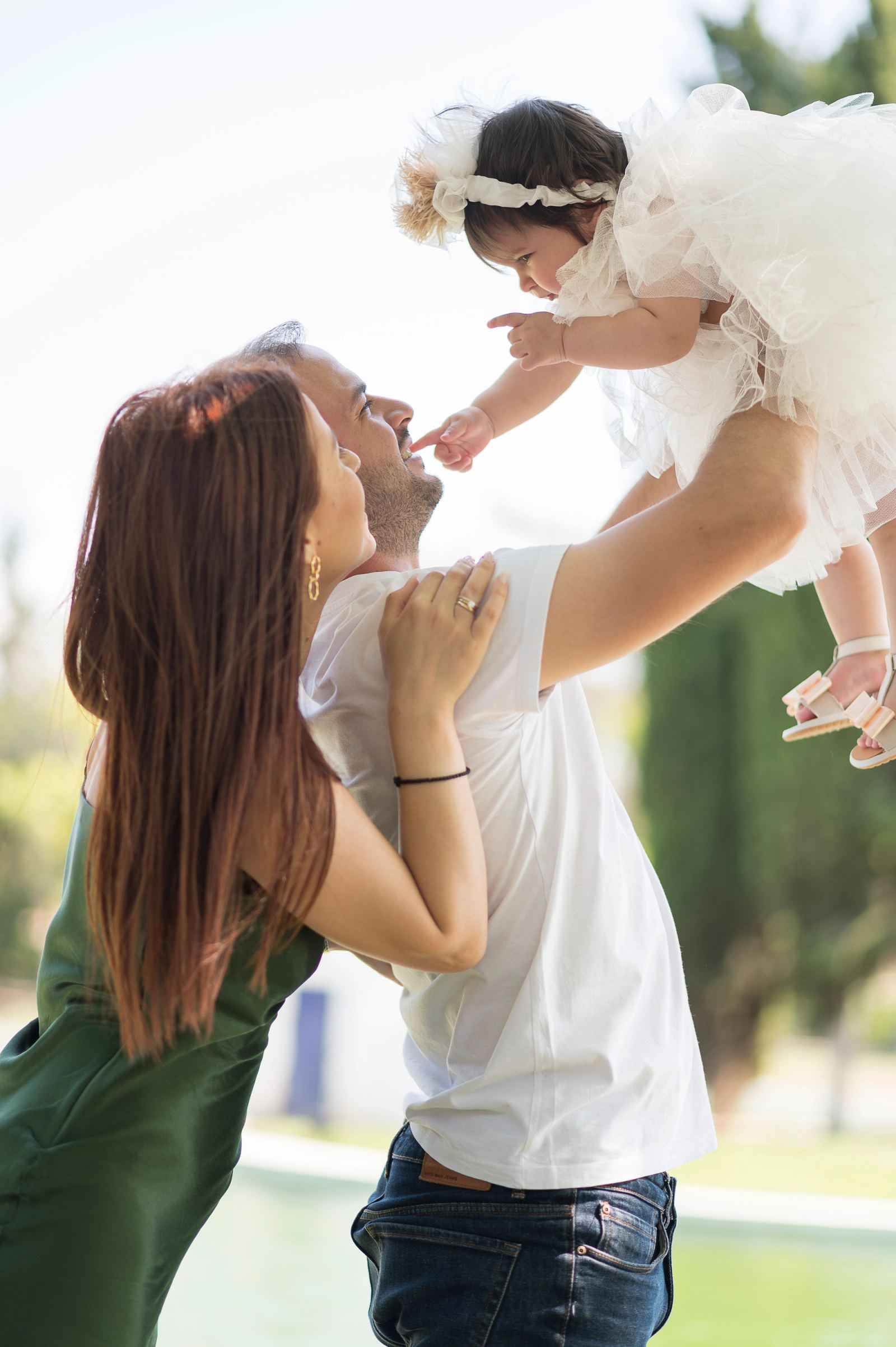 A man lifts the baby in a white dress high while a woman holds him in a garden.