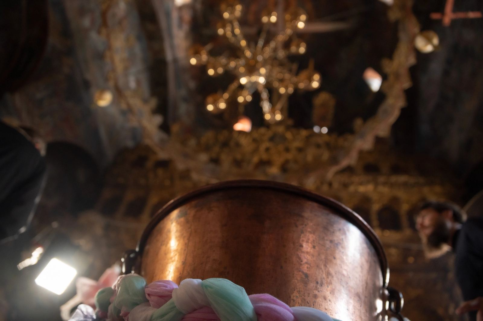 A copper font wrapped in a pink and green braid beneath a golden chandelier in a frescoed church.
