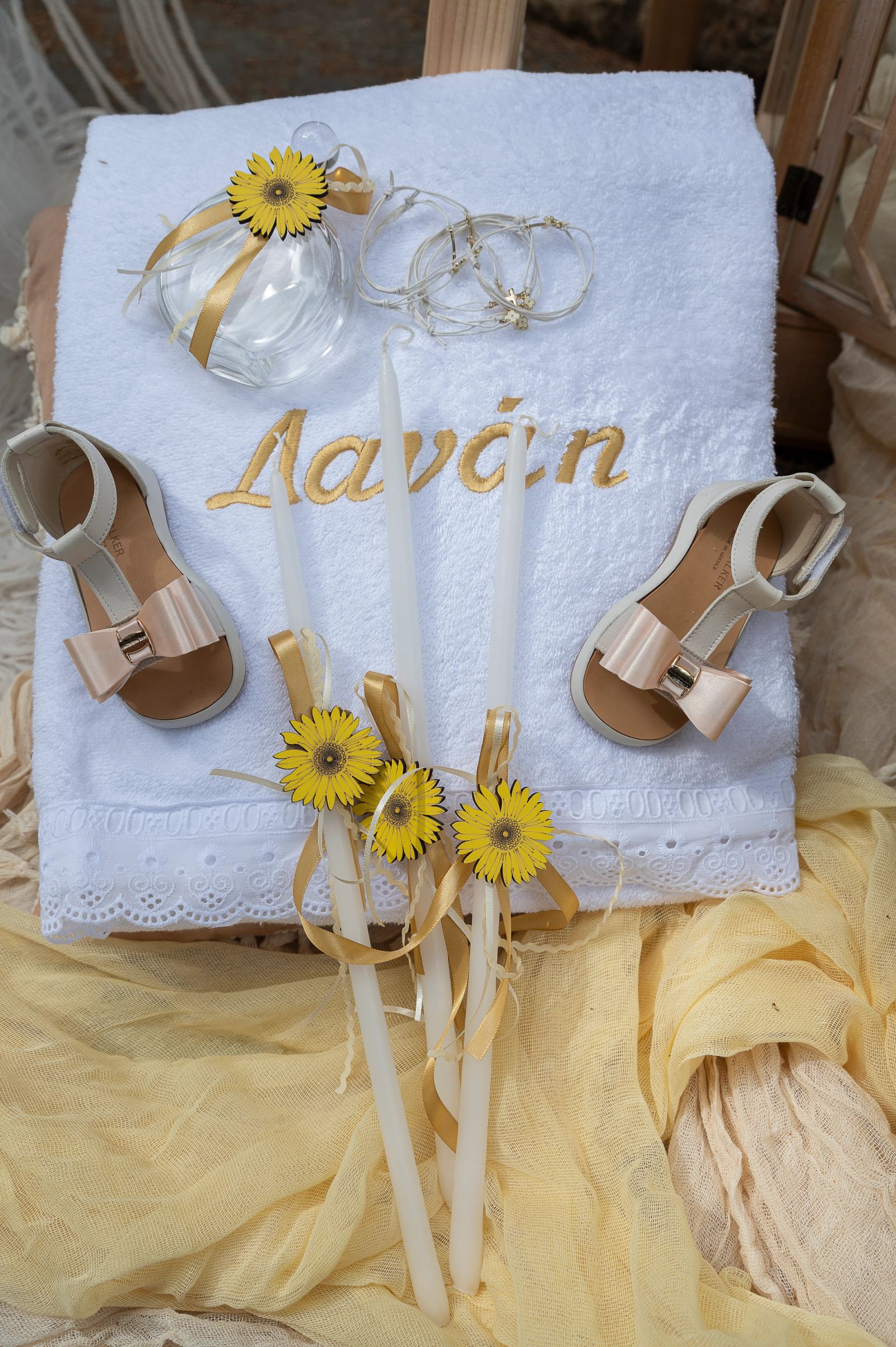 Embroidered white towel with sandals, candles with yellow daisies, a bottle and bracelets.