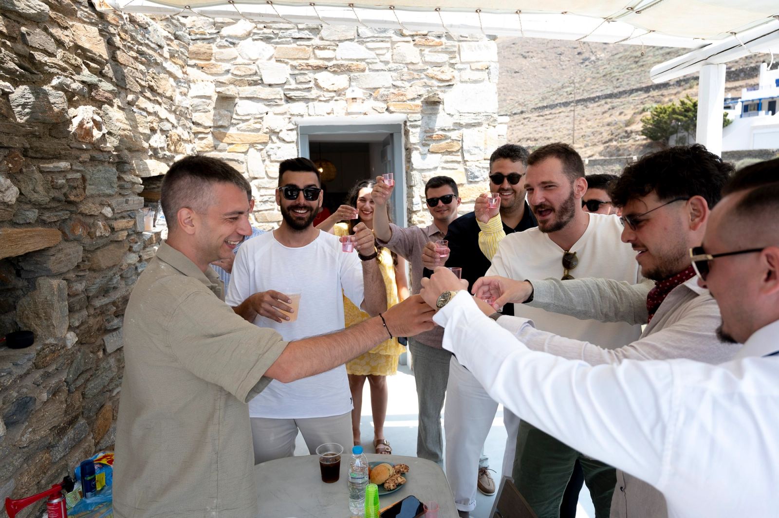 Ο γαμπρός και καλεσμένοι τσουγκρίζουν ποτηράκια σε αυλή με πέτρινους τοίχους και τέντα.