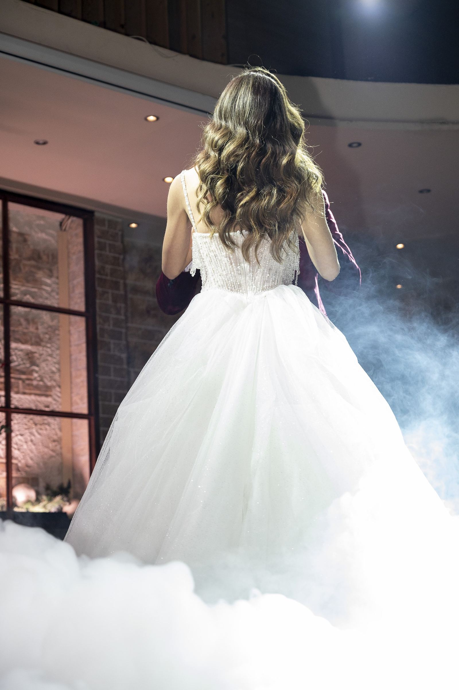 The bride seen from behind, with long wavy hair, dances in white fog in a hall with a stone wall.